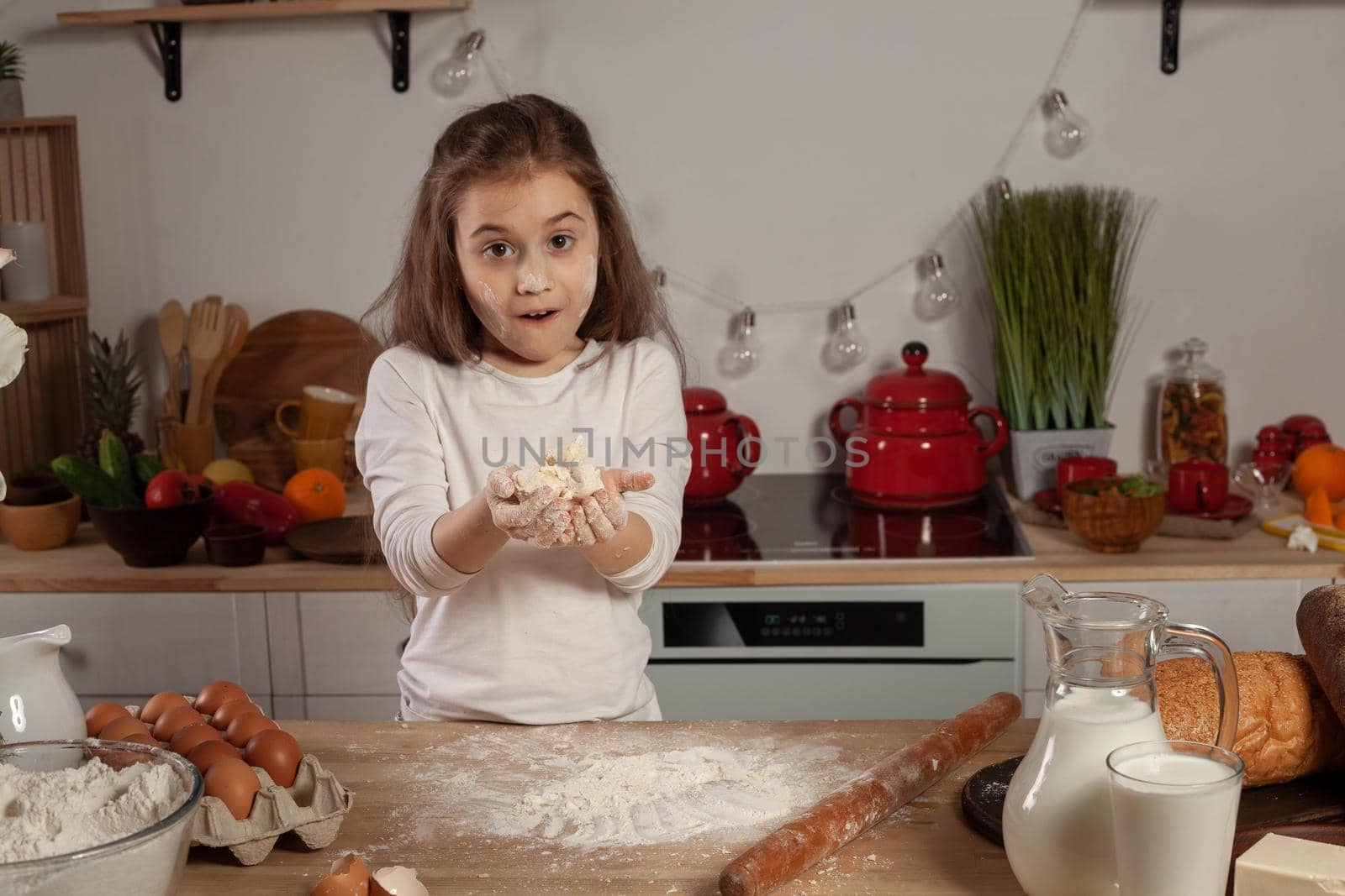 Charming little brunette lady dressed in a white blouse is showing a dough for baking a bread and looking at the camera at a kitchen against a white wall with shelves and bulbs on it. Happy childhood concept.