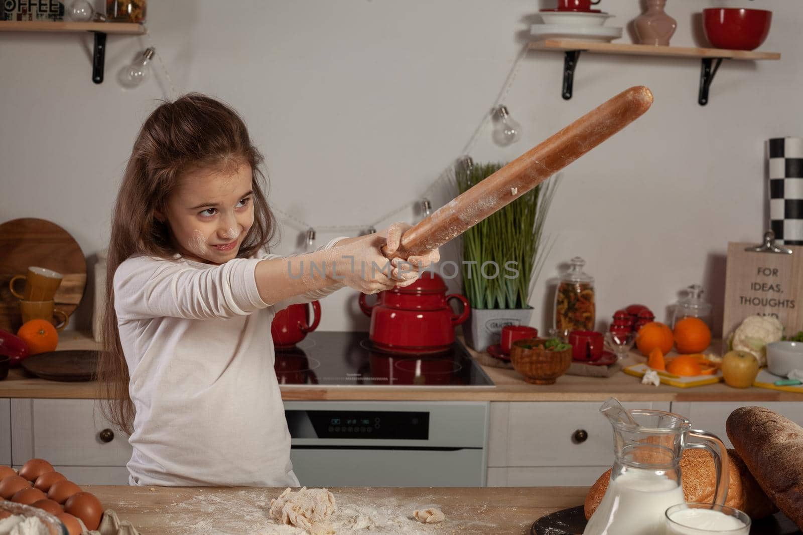 Beautiful little brunette lady dressed in a white blouse is playing with a rolling pin at a kitchen against a white wall with shelves and bulbs on it. Happy childhood concept.