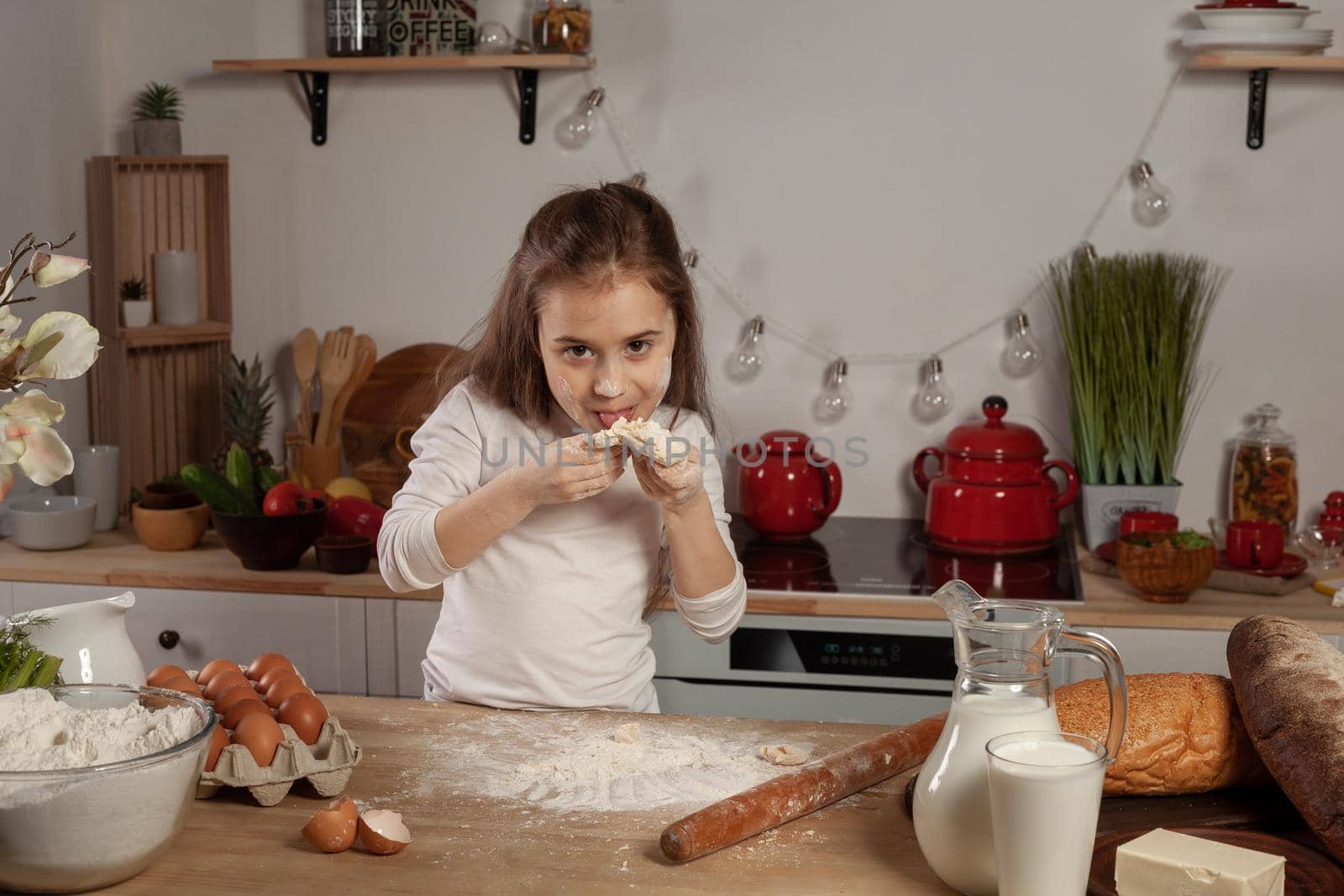 Charming little brunette child dressed in a white blouse is tasting the dough for baking a bread at a kitchen against a white wall with shelves and bulbs on it. Happy childhood concept.
