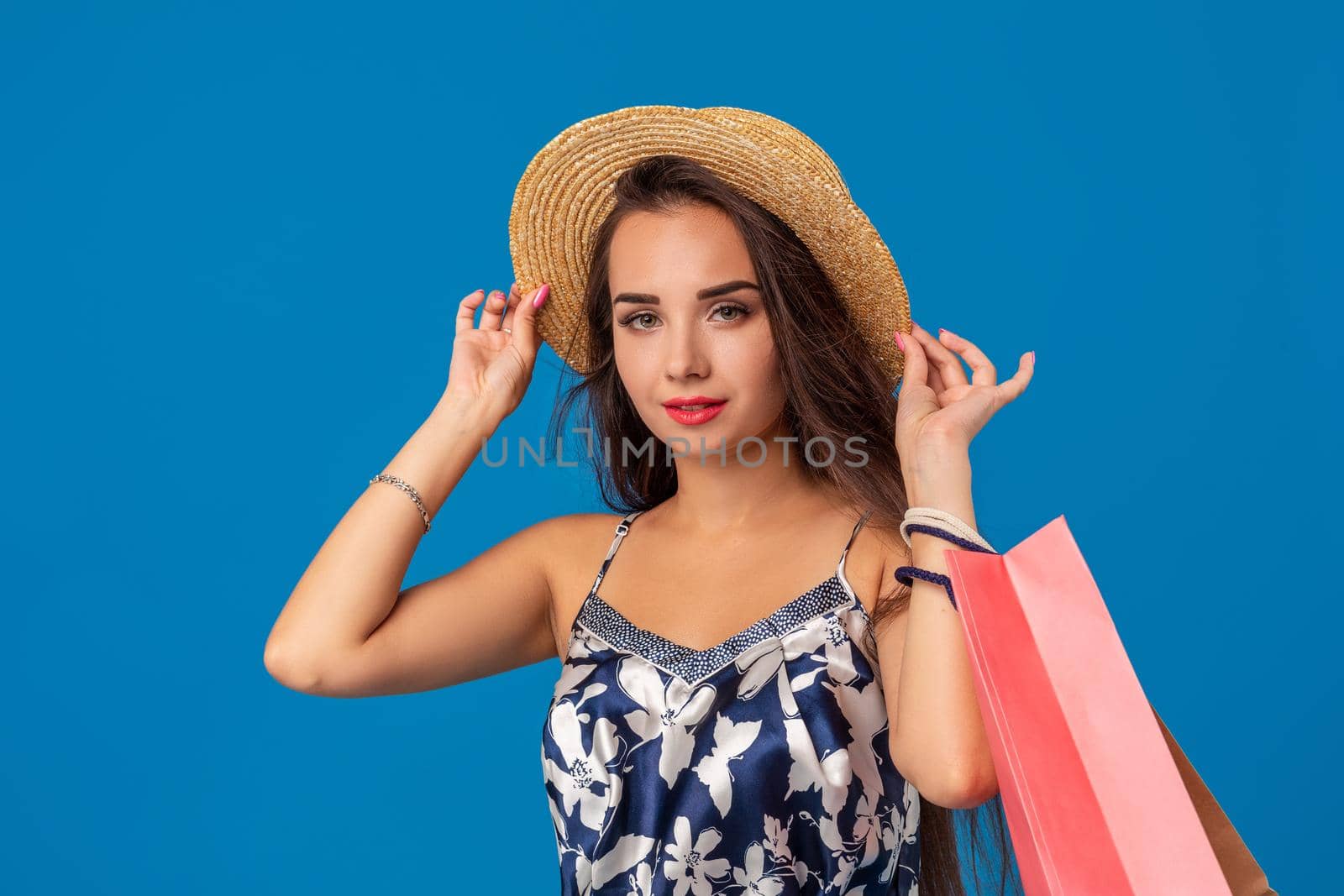 Portrait of a young woman in summer hat holding shopping bags and looking at the camera isolated over blue background. The concept of shopping