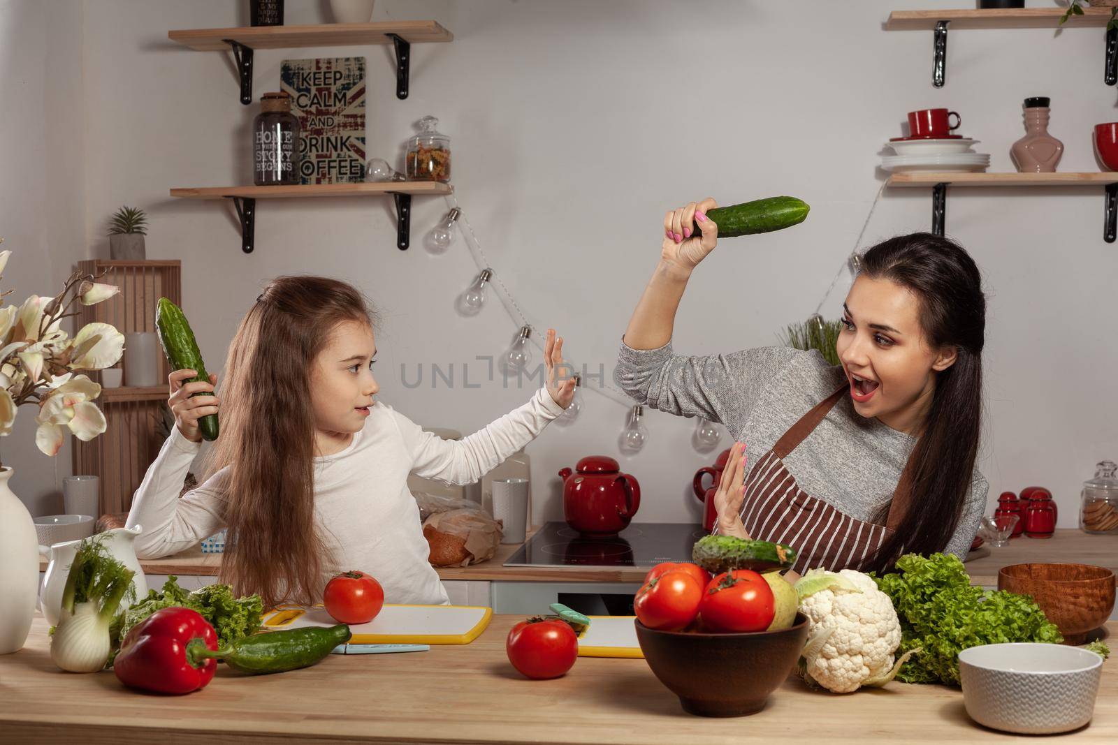 Happy loving family are cooking together. Wonderful mum and her child are fighting each other by cucumbers and having fun at the kitchen, against a white wall with shelves and bulbs on it. Homemade food and little helper.