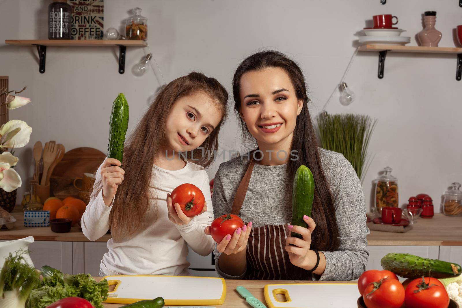 Happy loving family are cooking together. Good-looking mother and her daughter are posing with tomato and cucumber and having fun at the kitchen, against a white wall with shelves and bulbs on it. Homemade food and little helper.