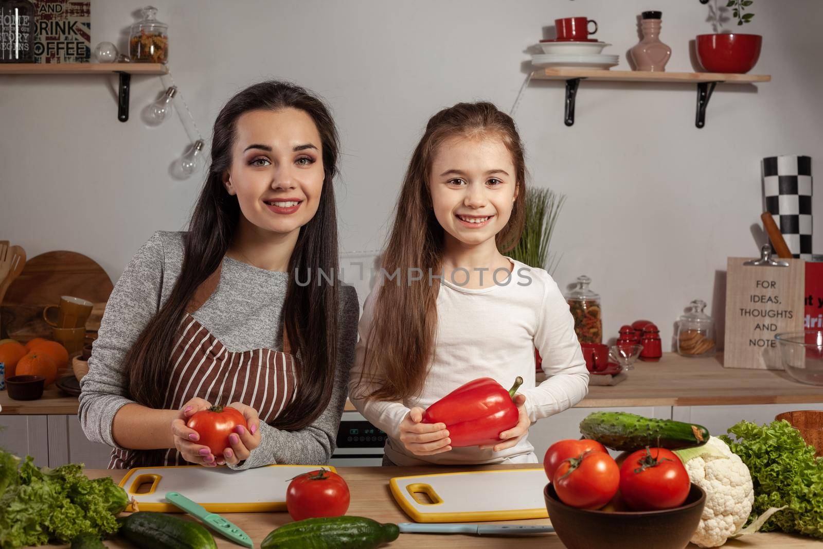 Happy loving family are cooking together. Loving mother and her daughter are making a vegetable salad and having fun at the kitchen, against a white wall with shelves and bulbs on it. Homemade food and little helper.