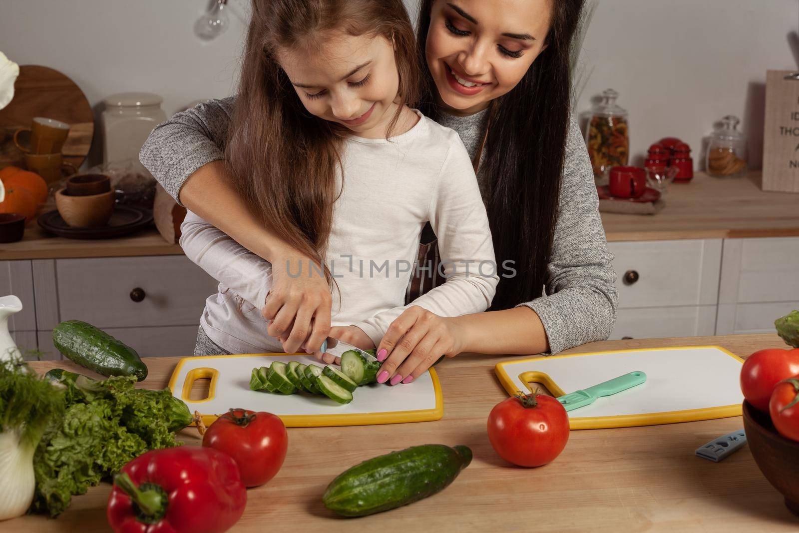 Happy loving family are cooking together. Elegant mother and her daughter are making a vegetable salad and cutting a cucumber at the kitchen, against a white wall with shelves and bulbs on it. Homemade food and little helper.