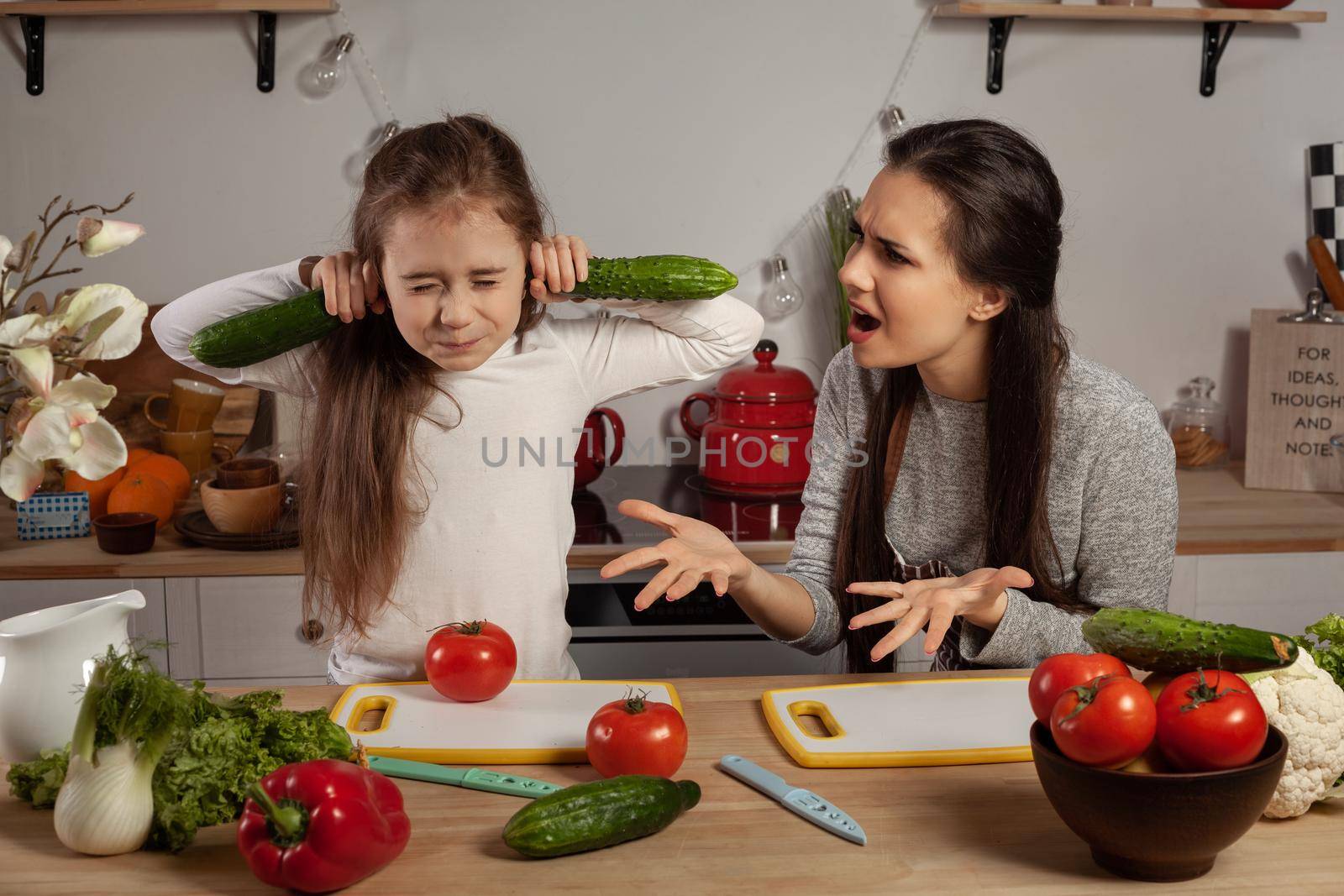 Happy loving family are cooking together. Brunette mother and her daughter are making a vegetable salad and quarrelling at the kitchen, against a white wall with shelves and bulbs on it. Homemade food and little helper.