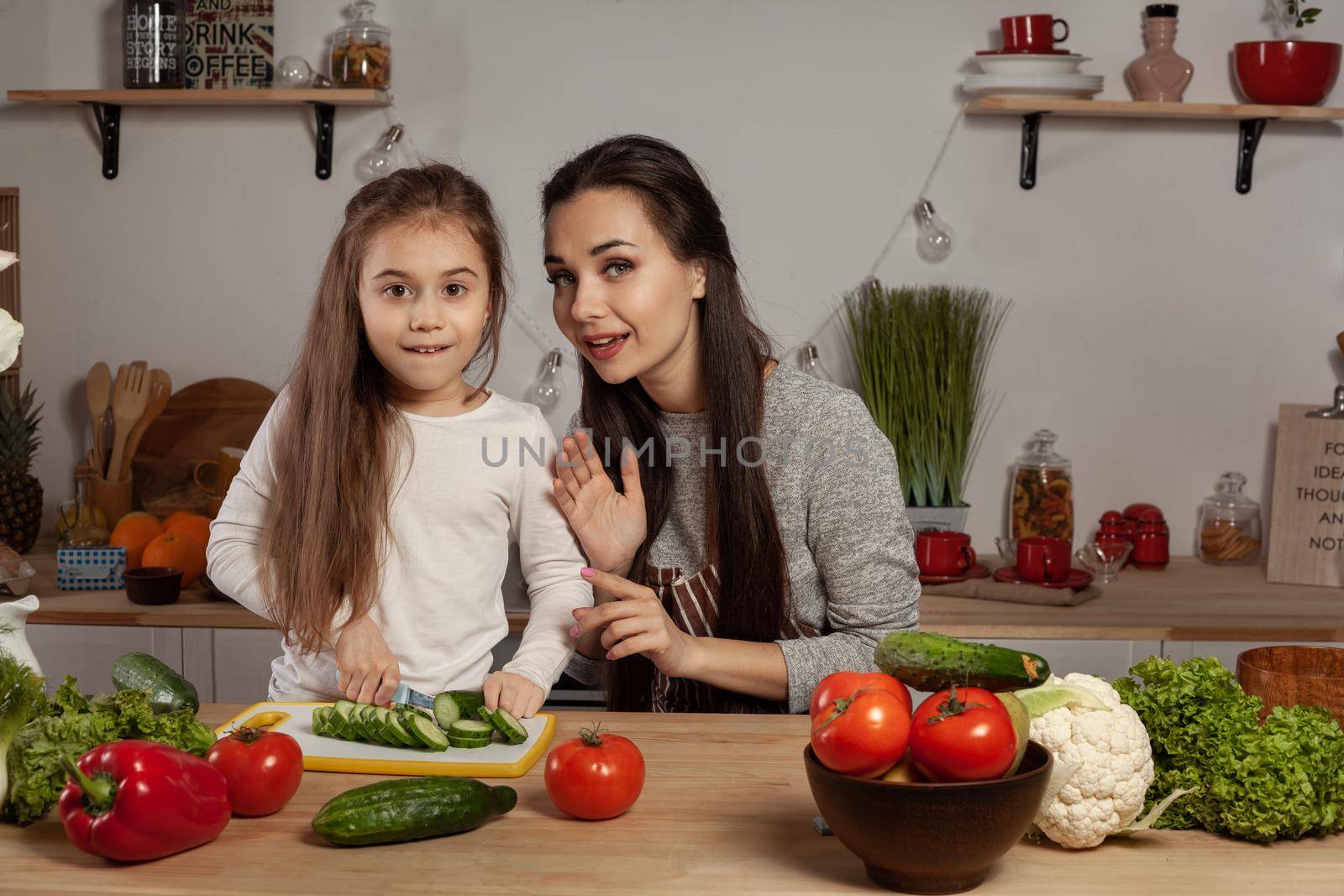 Happy loving family are cooking together. Cute mum and her child are making a vegetable salad and looking at the camera at the kitchen, against a white wall with shelves and bulbs on it. Homemade food and little helper.