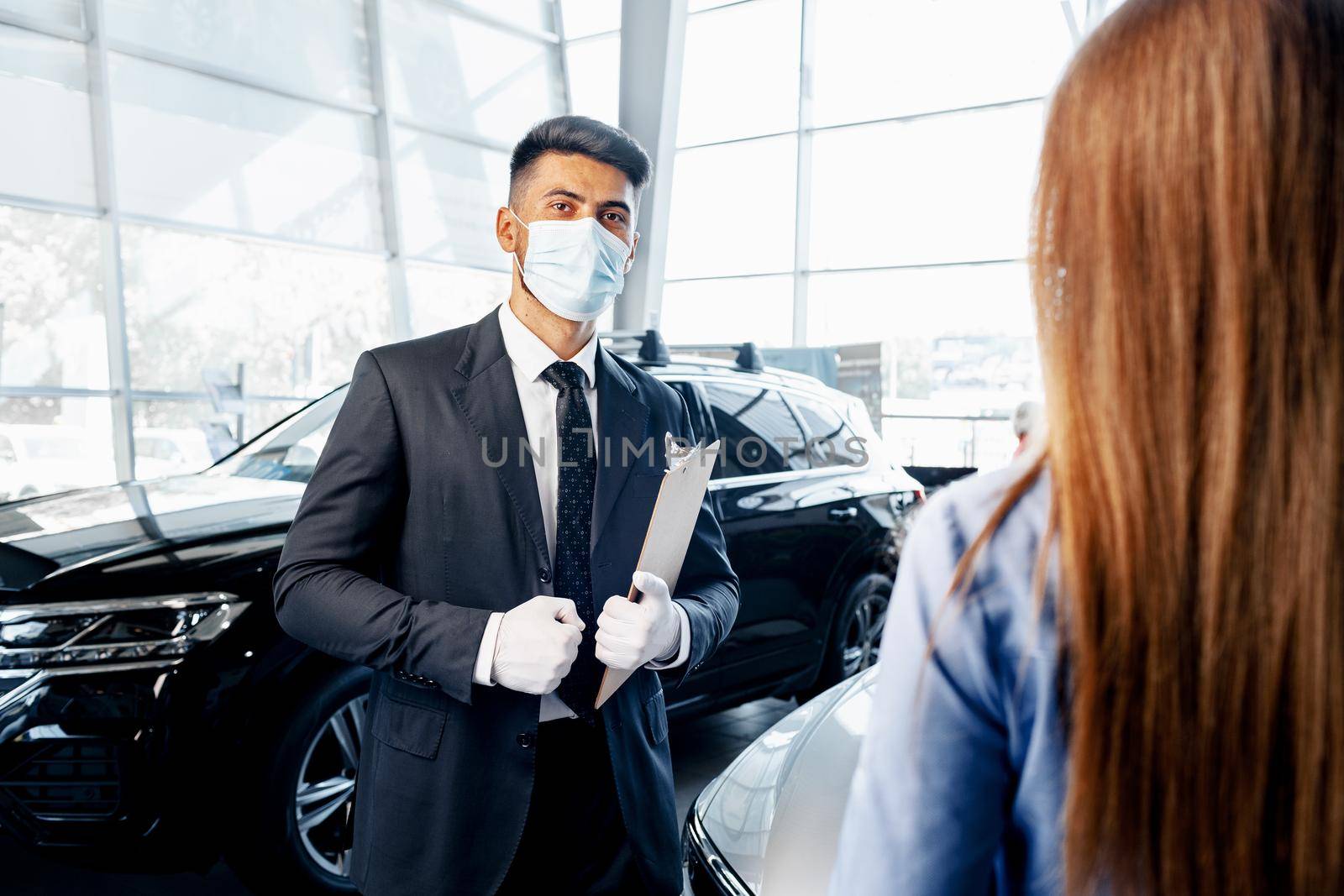 Man car salesman in face mask talking to a client in showroom by Fabrikasimf