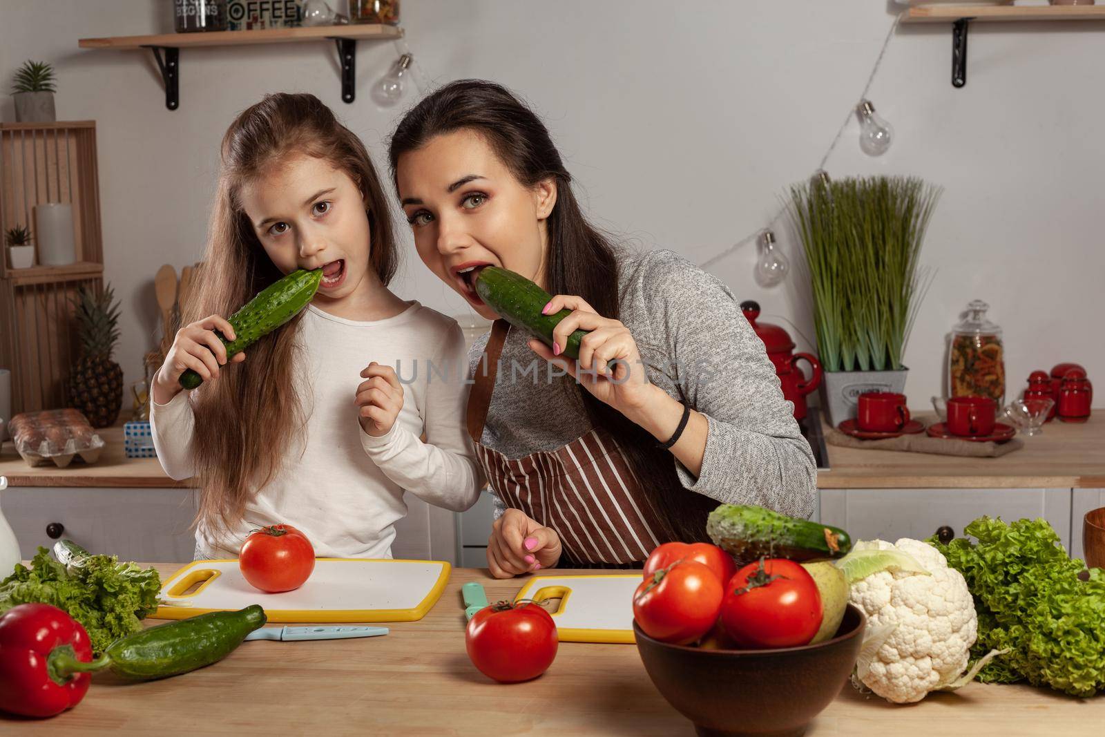 Happy loving family are cooking together. Good-looking mommy and her little girl are posing with tomato and cucumber and having fun at the kitchen, against a white wall with shelves and bulbs on it. Homemade food and little helper.