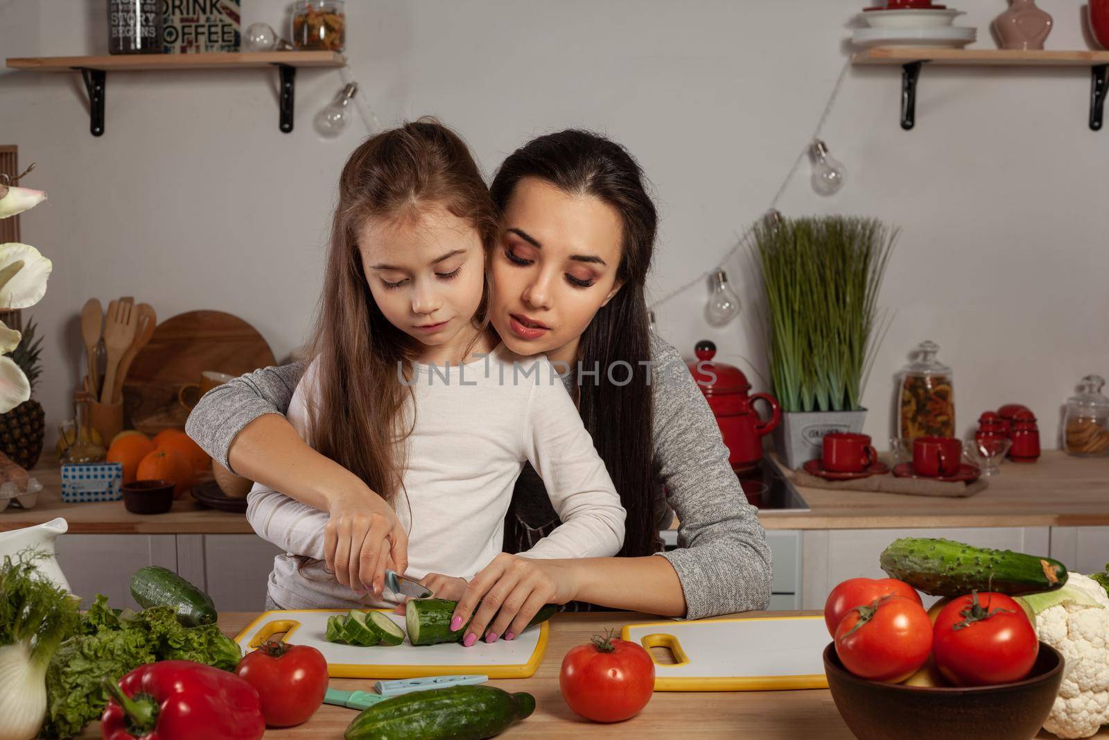 Happy loving family are cooking together. Elegant mommy and her little girl are making a vegetable salad and cutting a cucumber at the kitchen, against a white wall with shelves and bulbs on it. Homemade food and little helper.