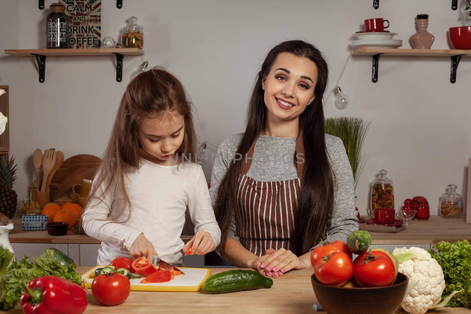 Happy loving family are cooking together. Charming mother and her little princess are making a vegetable salad, smiling and having fun at the kitchen, against a white wall with shelves and bulbs on it. Homemade food and little helper.