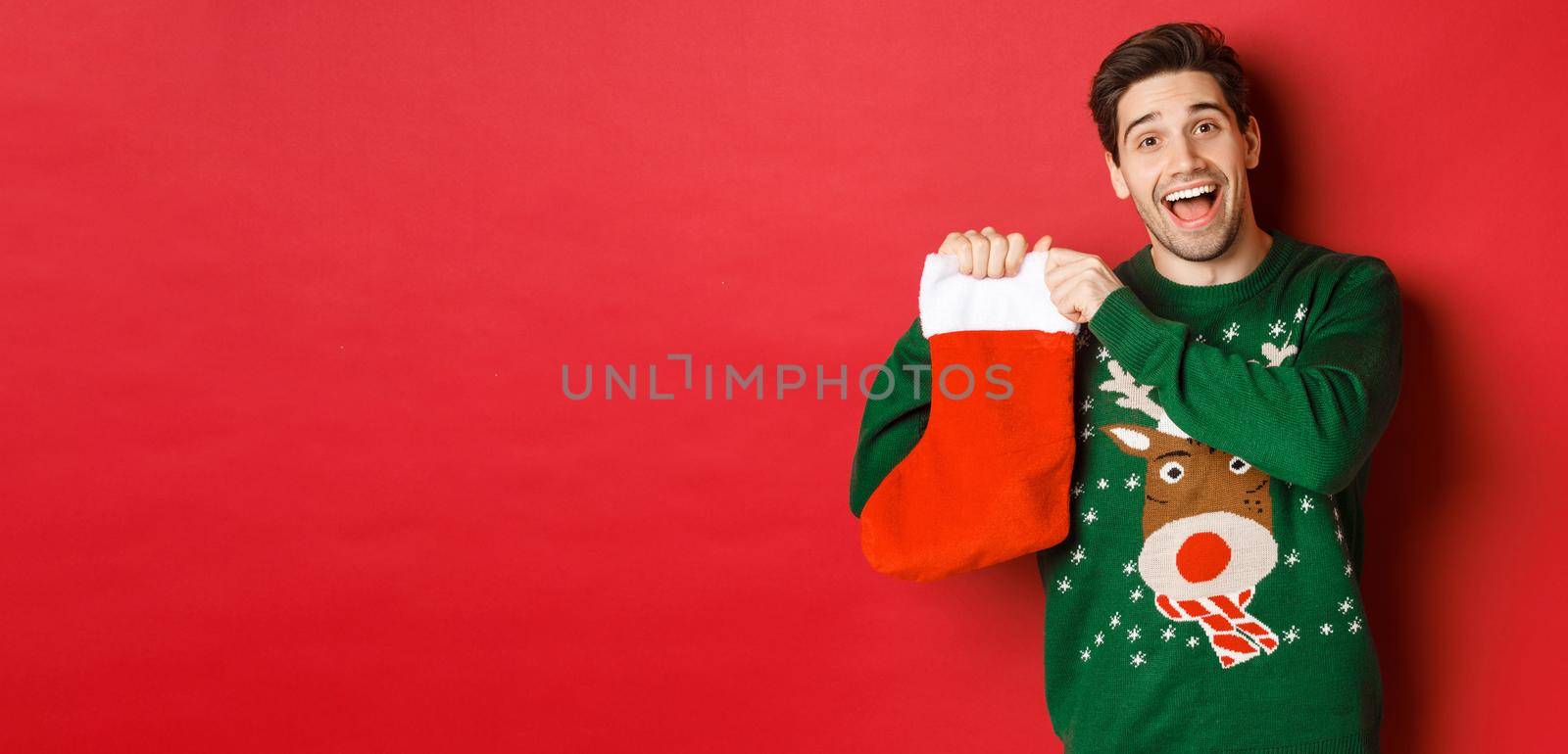 Image of attractive cheerful man in sweater, holding christmas stocking with gifts, celebrating winter holidays, standing over red background.