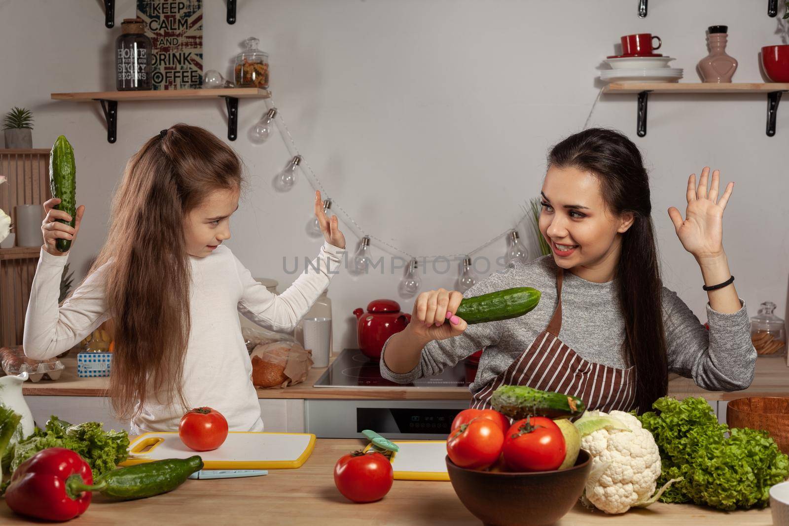 Happy loving family are cooking together. Wonderful mom and her kid are fighting each other by cucumbers and having fun at the kitchen, against a white wall with shelves and bulbs on it. Homemade food and little helper.