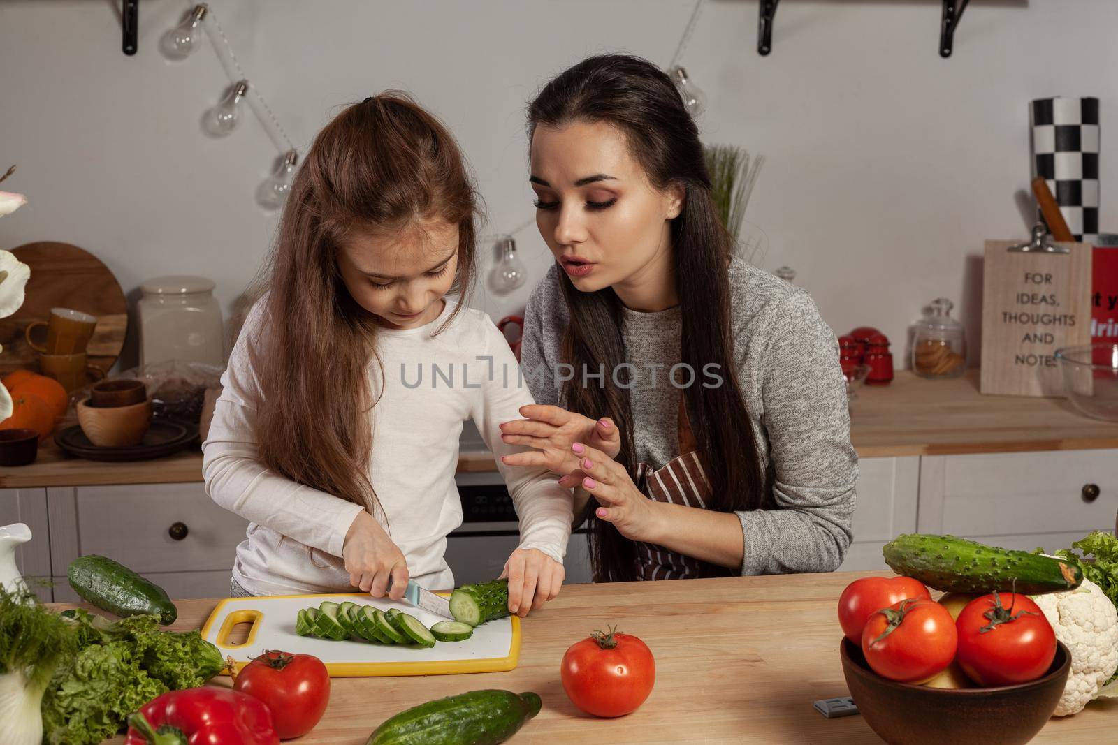 Happy loving family are cooking together. Cute mother and her little princess are making a vegetable salad and cutting a cucumber at the kitchen, against a white wall with shelves and bulbs on it. Homemade food and little helper.