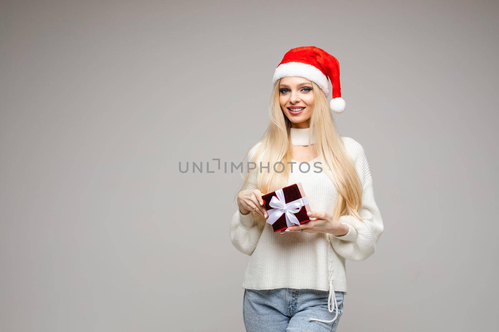 Portrait of happy pretty female wearing Santa hat while posing with gift, isolated on grey background. New Year holiday concept