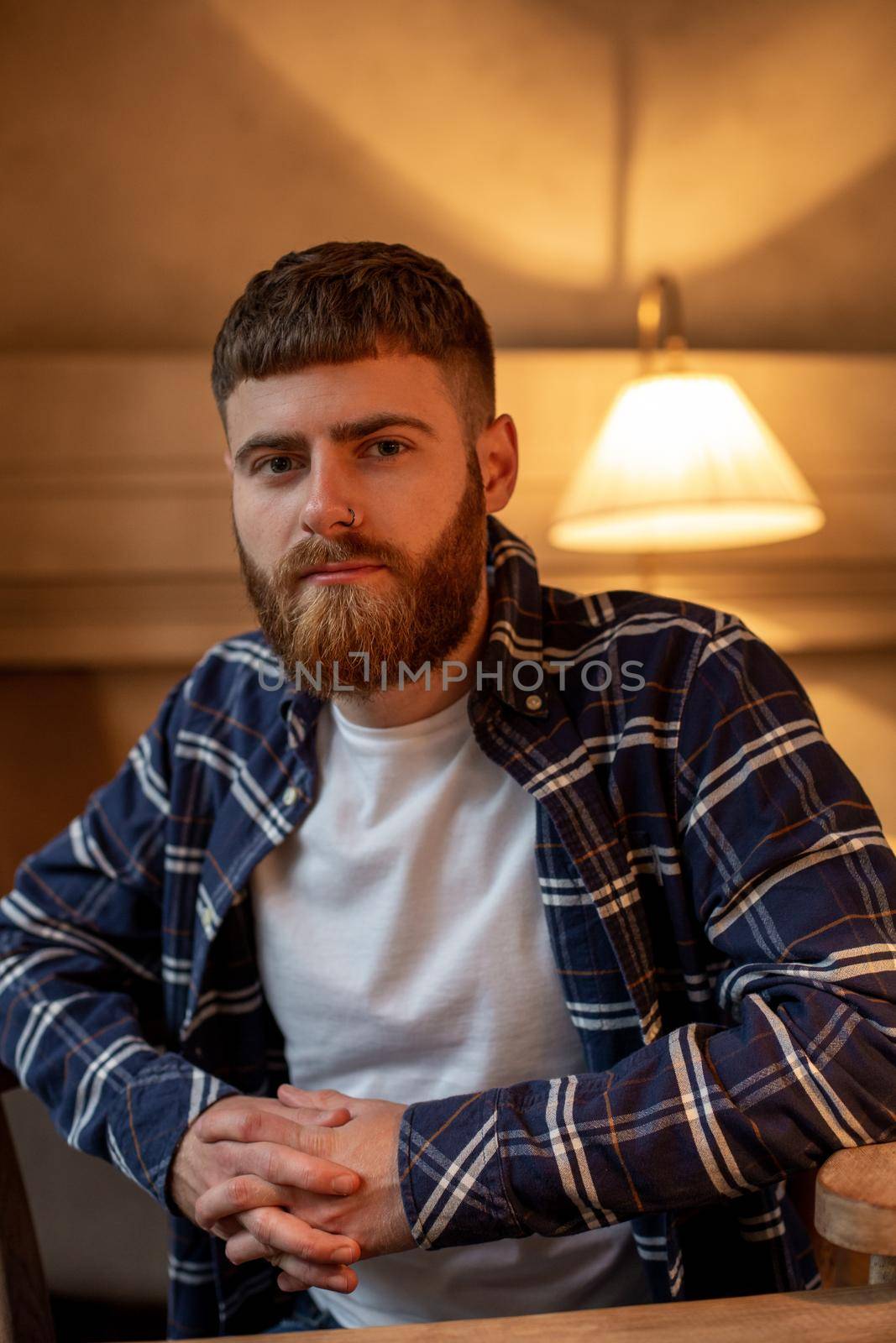 Portrait handsome bearded man wearing plaid shirt at modern cafe. Guy sitting in wooden chair and relaxing.
