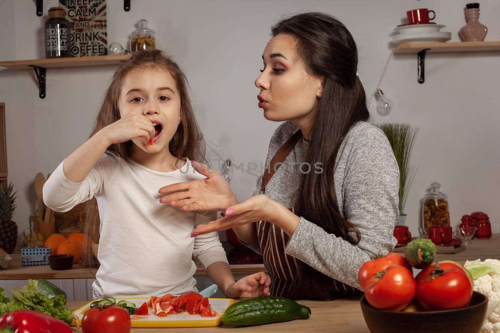 Happy loving family are cooking together. Beautiful mommy and her little girl are making a vegetable salad and tasting it at the kitchen, against a white wall with shelves and bulbs on it. Homemade food and little helper.