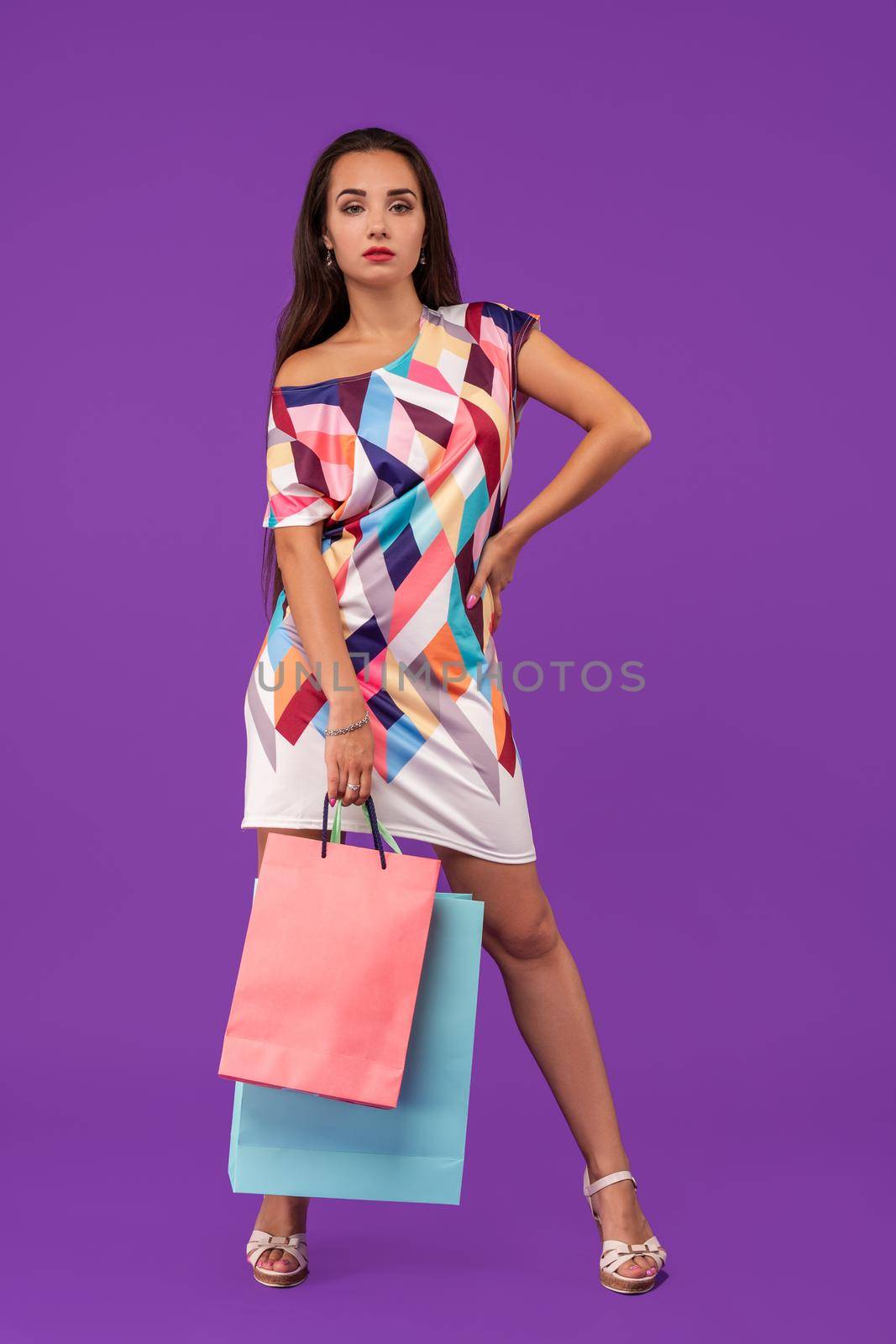 In love with shopping. Beautiful young woman holding shopping bags and looking at the camera while standing against purple background. Full-height image.