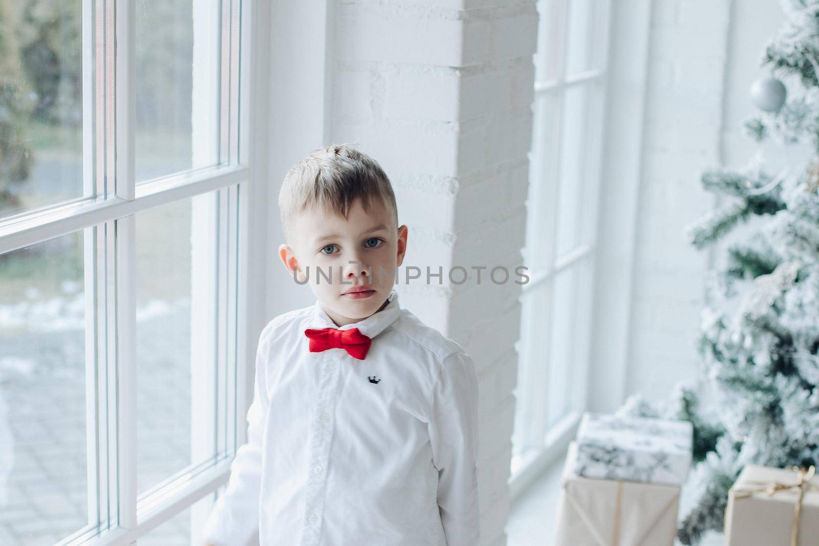 little caucasian boy sits near the christmas tree with a gift by StudioLucky