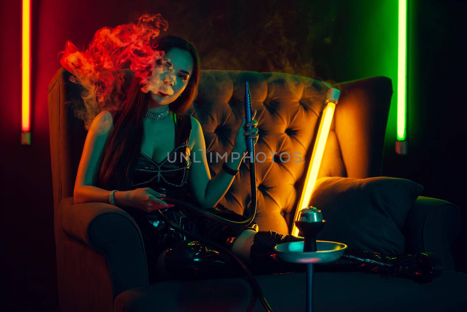 Amazing brunette female with a perfect make-up, in a sparkling necklace, leather dress and patent boots is smoking a hookah exhaling a smoke and looking at the camera sitting on an arm-chair at a luxury night club. Concept of pleasant spending time. Dark background with colorful neon backlights.