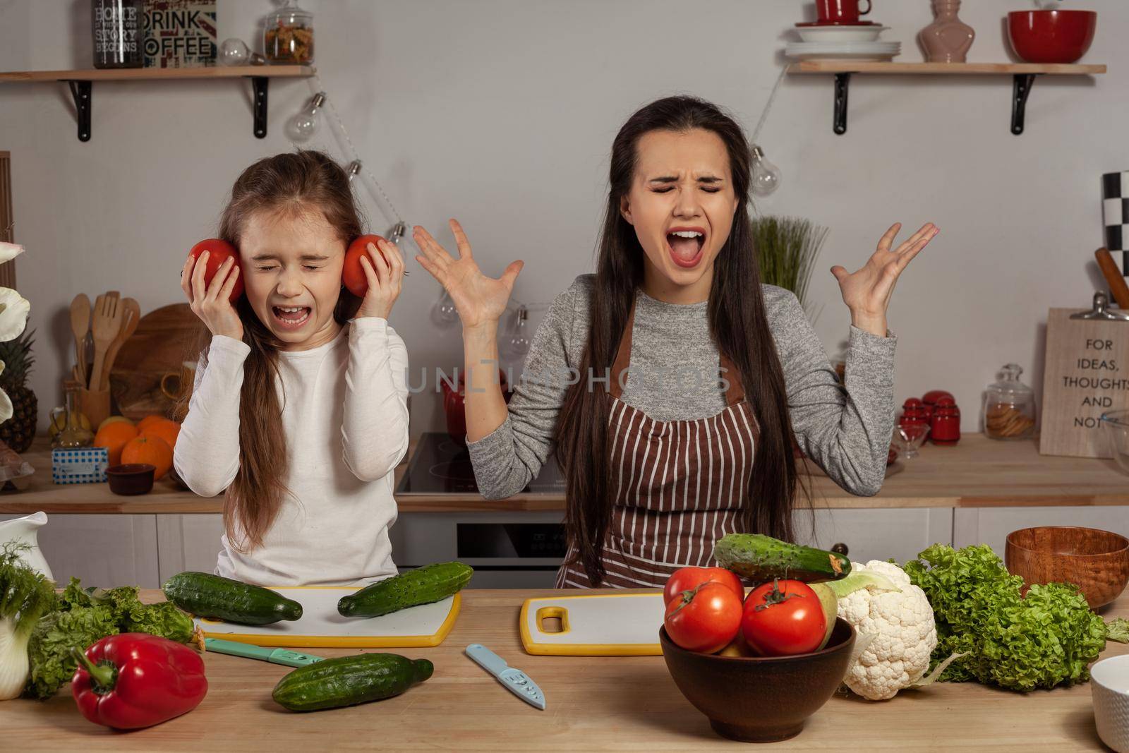 Happy loving family are cooking together. Elegant mother and her little princess are making a vegetable salad and quarrelling at the kitchen, against a white wall with shelves and bulbs on it. Homemade food and little helper.