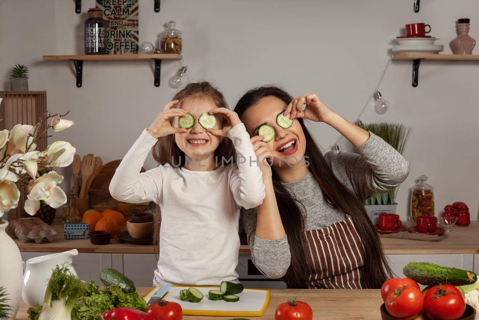 Happy loving family are cooking together. Alluring mom and her kid are making a vegetable salad and fooling around at the kitchen, against a white wall with shelves and bulbs on it. Homemade food and little helper.