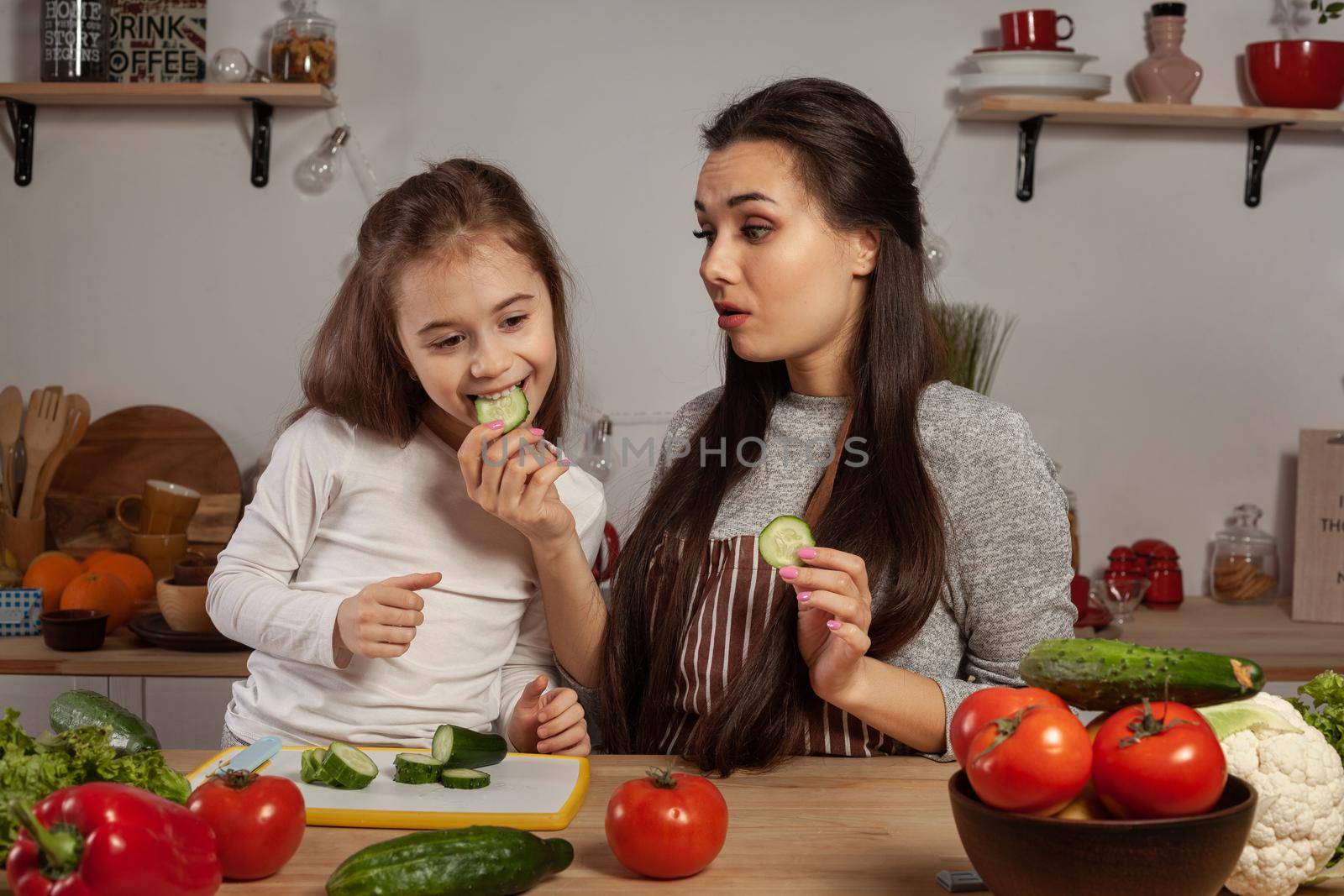Happy loving family are cooking together. Attractive mommy and her little girl are making a vegetable salad and tasting a cucumber at the kitchen, against a white wall with shelves and bulbs on it. Homemade food and little helper.