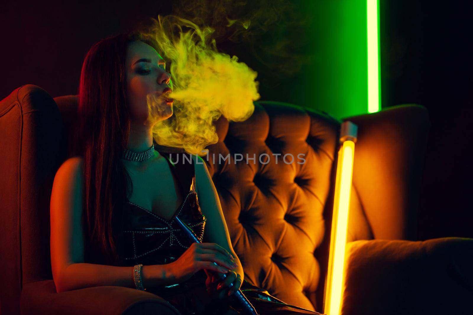 Close-up shot of a pretty brunette female with a perfect make-up, in a sparkling necklace and a leather dress smoking a hookah exhaling a smoke sitting sideways on an arm-chair at a luxury night club. Concept of pleasant spending time. Dark background with colorful neon backlights.