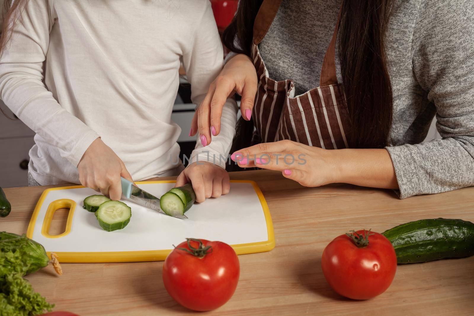 Close-up shot of a happy loving family cooking together. Mother and her daughter are making a vegetable salad and cutting a cucumber at the kitchen. Homemade food and little helper.