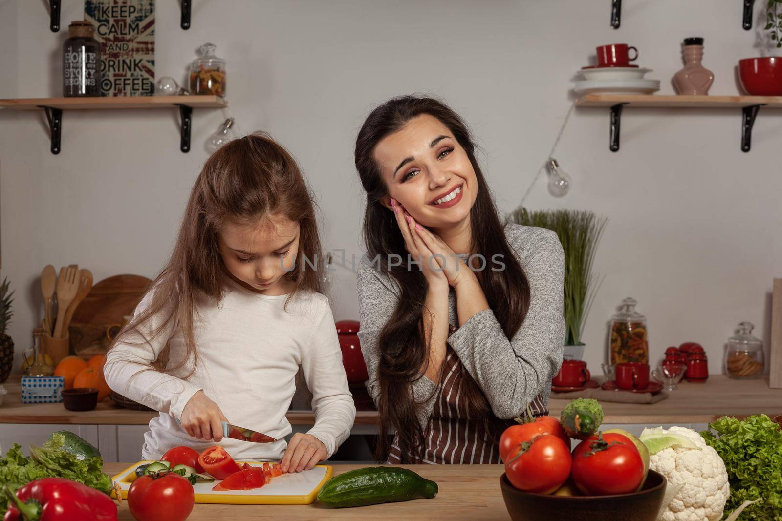 Happy loving family are cooking together. Gorgeous mother and her daughter are making a vegetable salad and mommy is touched by her offspring at the kitchen, against a white wall with shelves and bulbs on it. Homemade food and little helper.