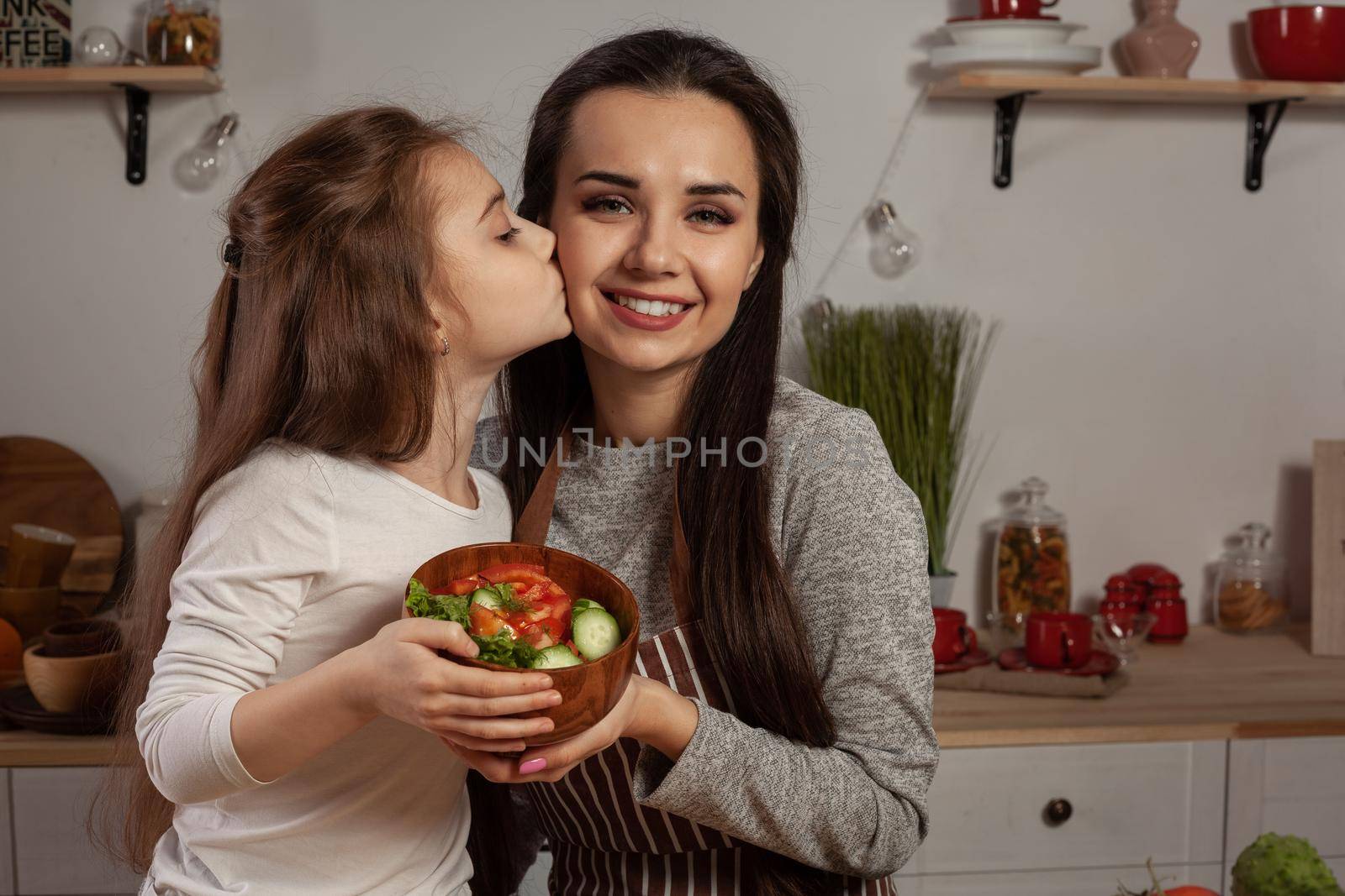 Happy loving family are cooking together. Smiling mother and her daughter are making a vegetable salad, kissing and smiling at the kitchen, against a white wall with shelves and bulbs on it. Homemade food and little helper.