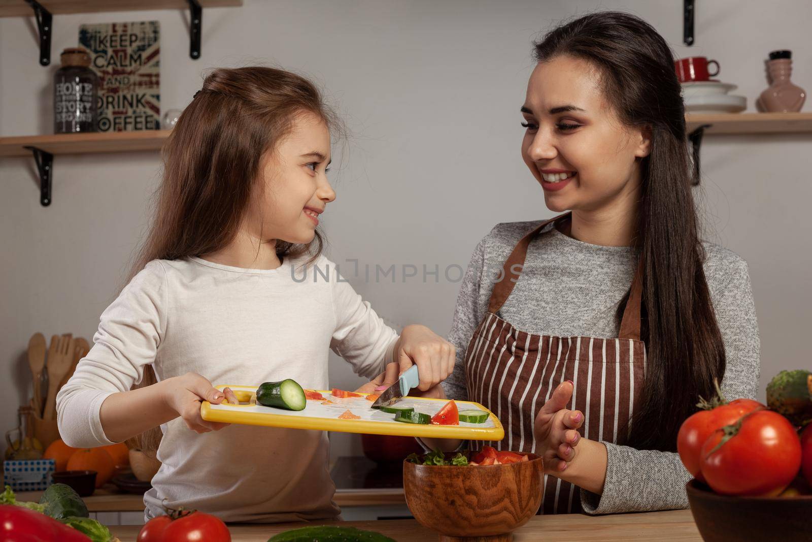 Happy loving family are cooking together. Beautiful mother and her daughter are making a vegetable salad and looking at each other at the kitchen, against a white wall with shelves and bulbs on it. Homemade food and little helper.