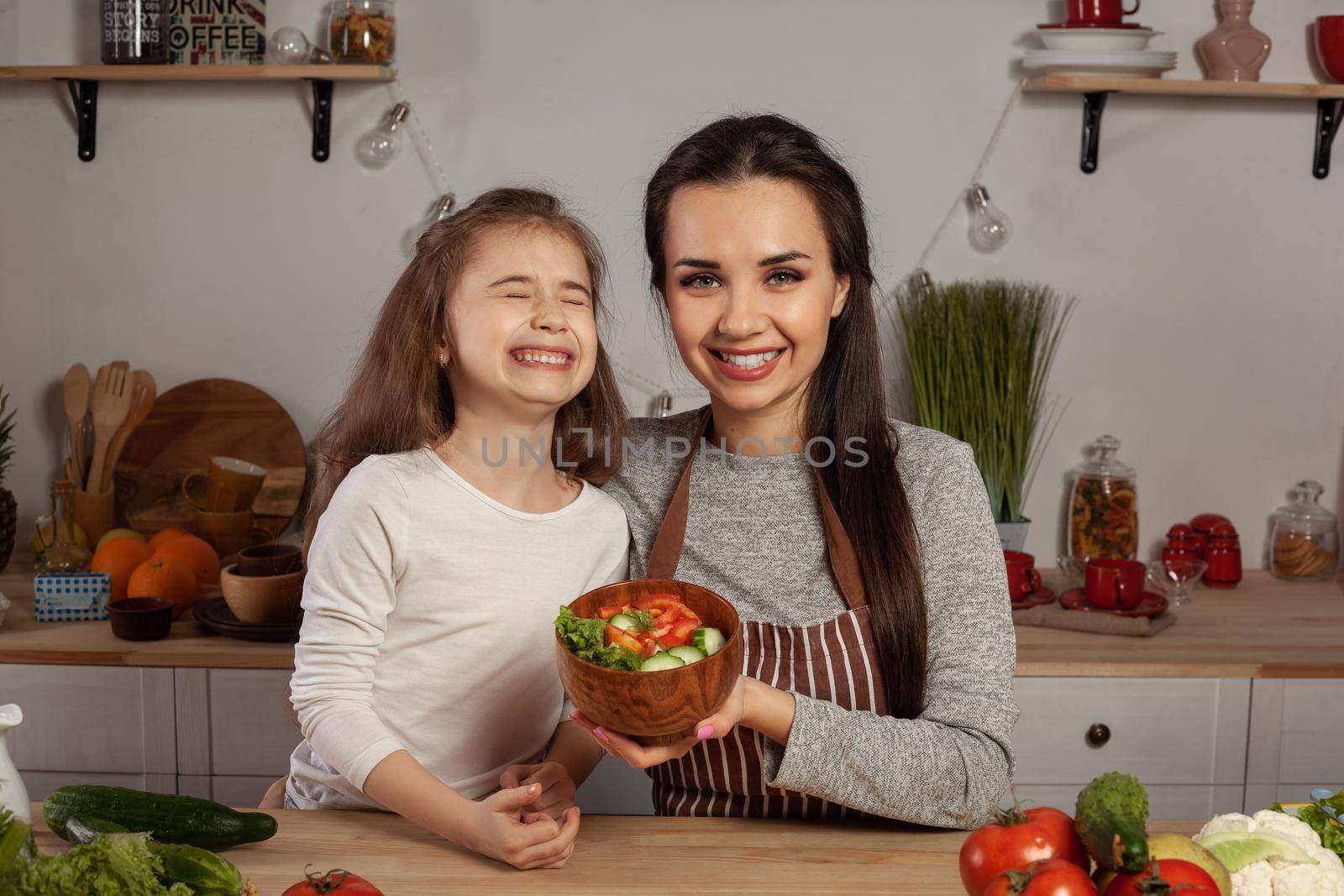 Happy loving family are cooking together. Smiling mother and her little princess are making a vegetable salad and laughing at the kitchen, against a white wall with shelves and bulbs on it. Homemade food and little helper.