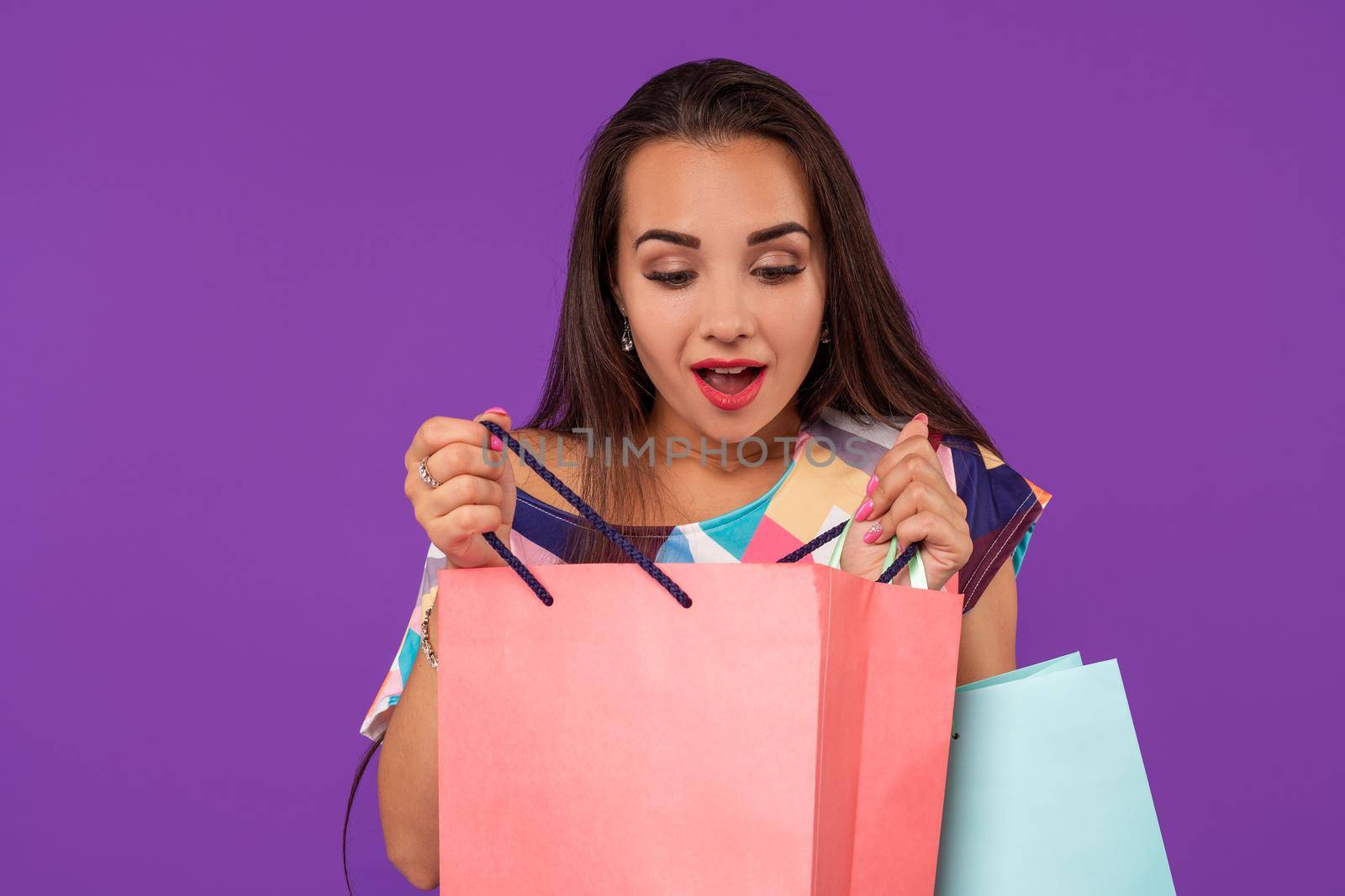 Beautiful emotional woman peeks into a package, in the hands of multi-colored shopping bags on a purple background. Studio shot. Copy space