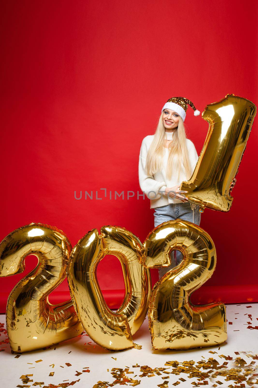 Xmas smiling young girl in santa hat with golden balloons in 2021 shape on red background for christmas holiday design by StudioLucky