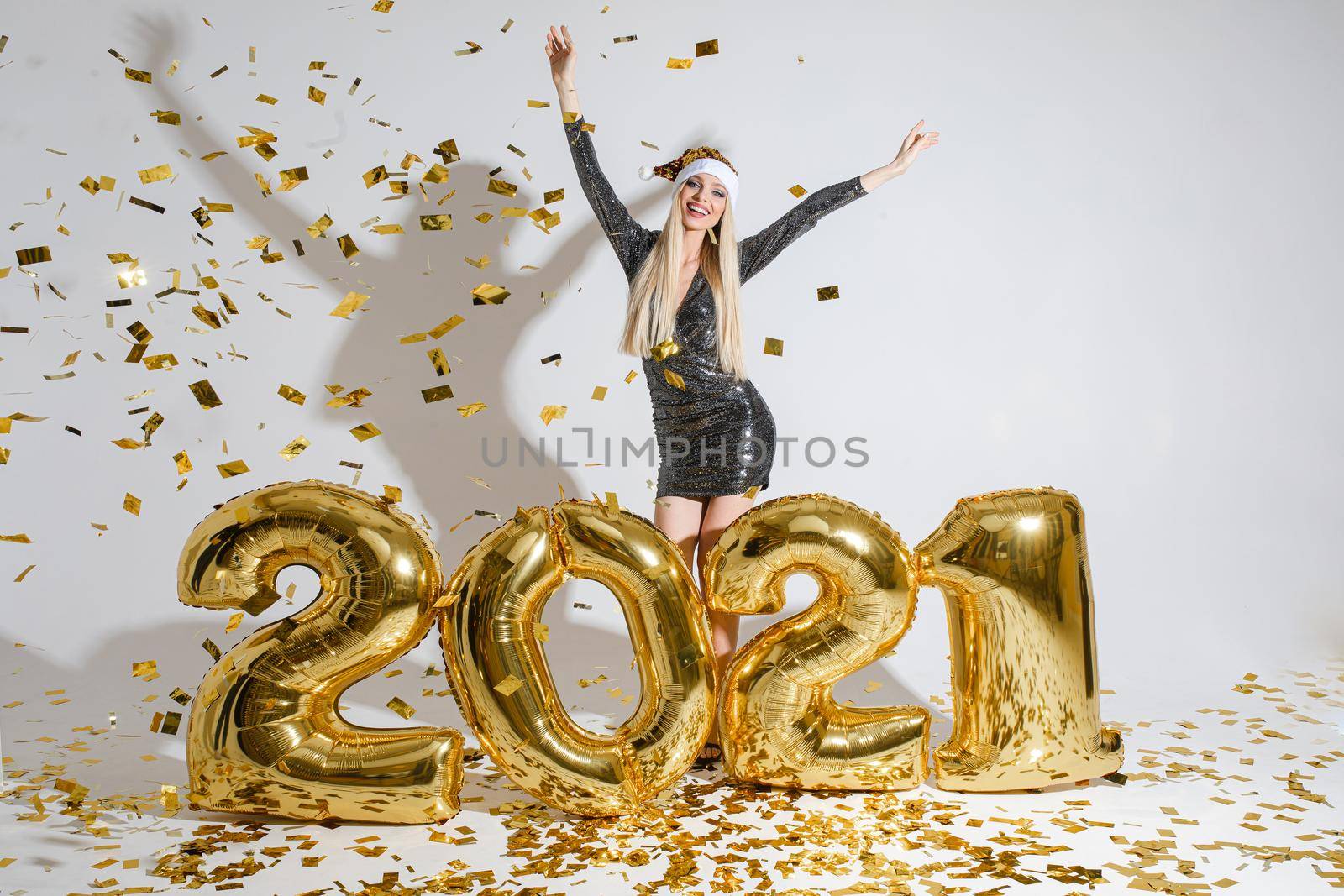 Stock photo of happy blonde girl in cocktail dress and Santa hat celebrating New Year with raised arms