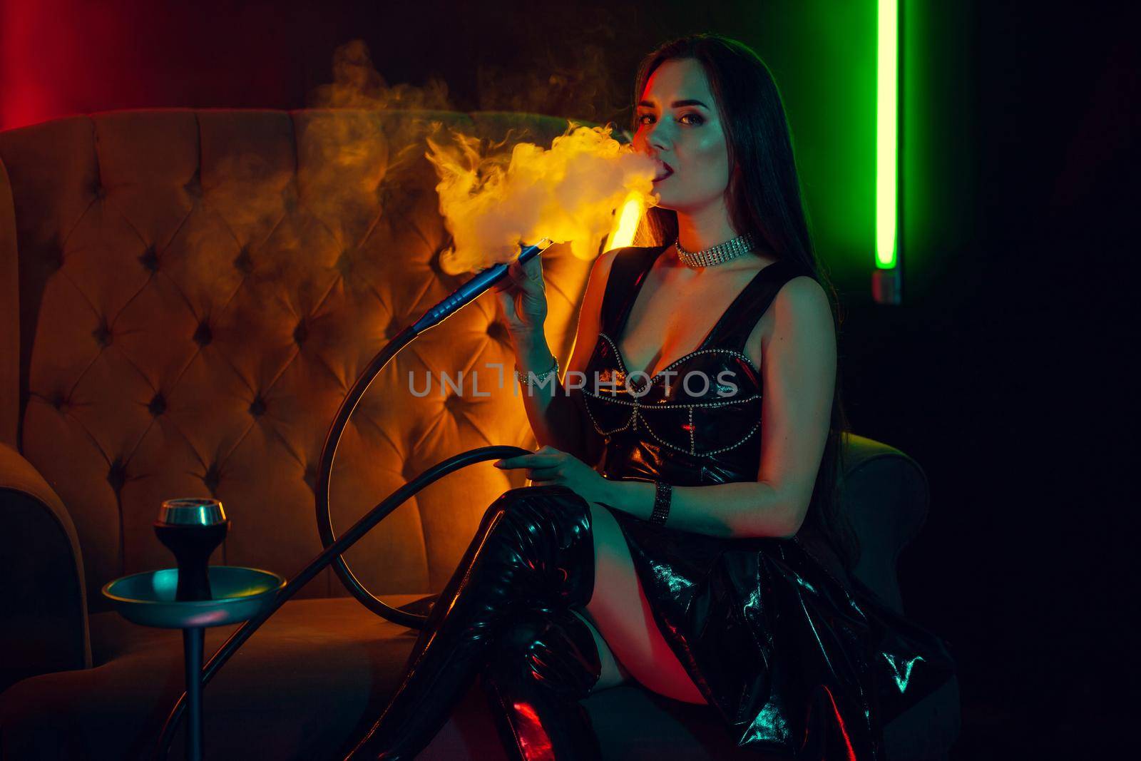 Beautiful brunette woman with a perfect make-up, in a sparkling necklace, leather dress and patent boots is smoking a hookah exhaling a smoke sitting sideways on an arm-chair at a luxury night club. Concept of pleasant spending time. Dark background with colorful neon backlights.