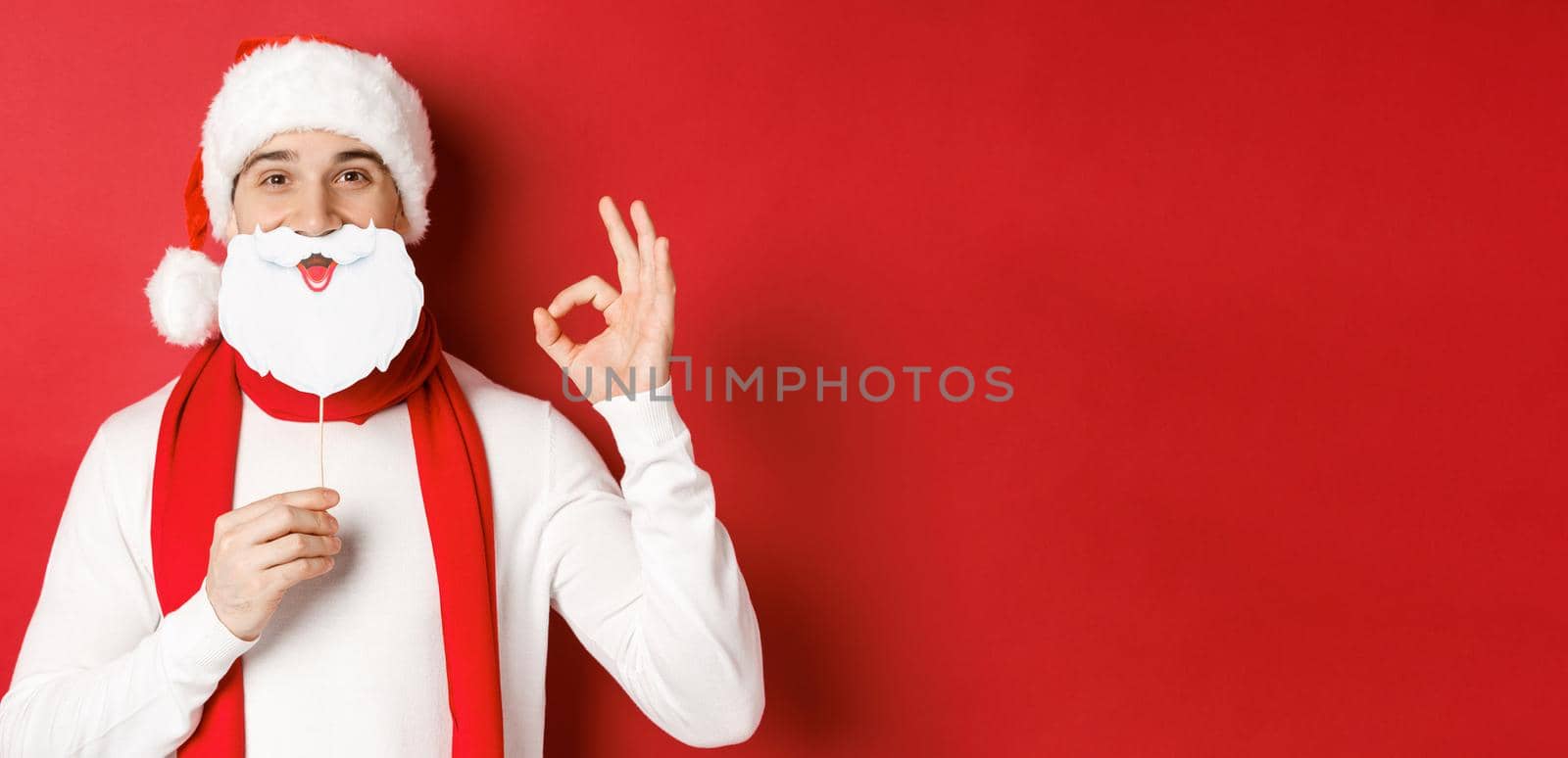 Concept of christmas, winter holidays and celebration. Pleased handsome man in santa hat, holding long white beard mask and showing okay sign, standing over red background.