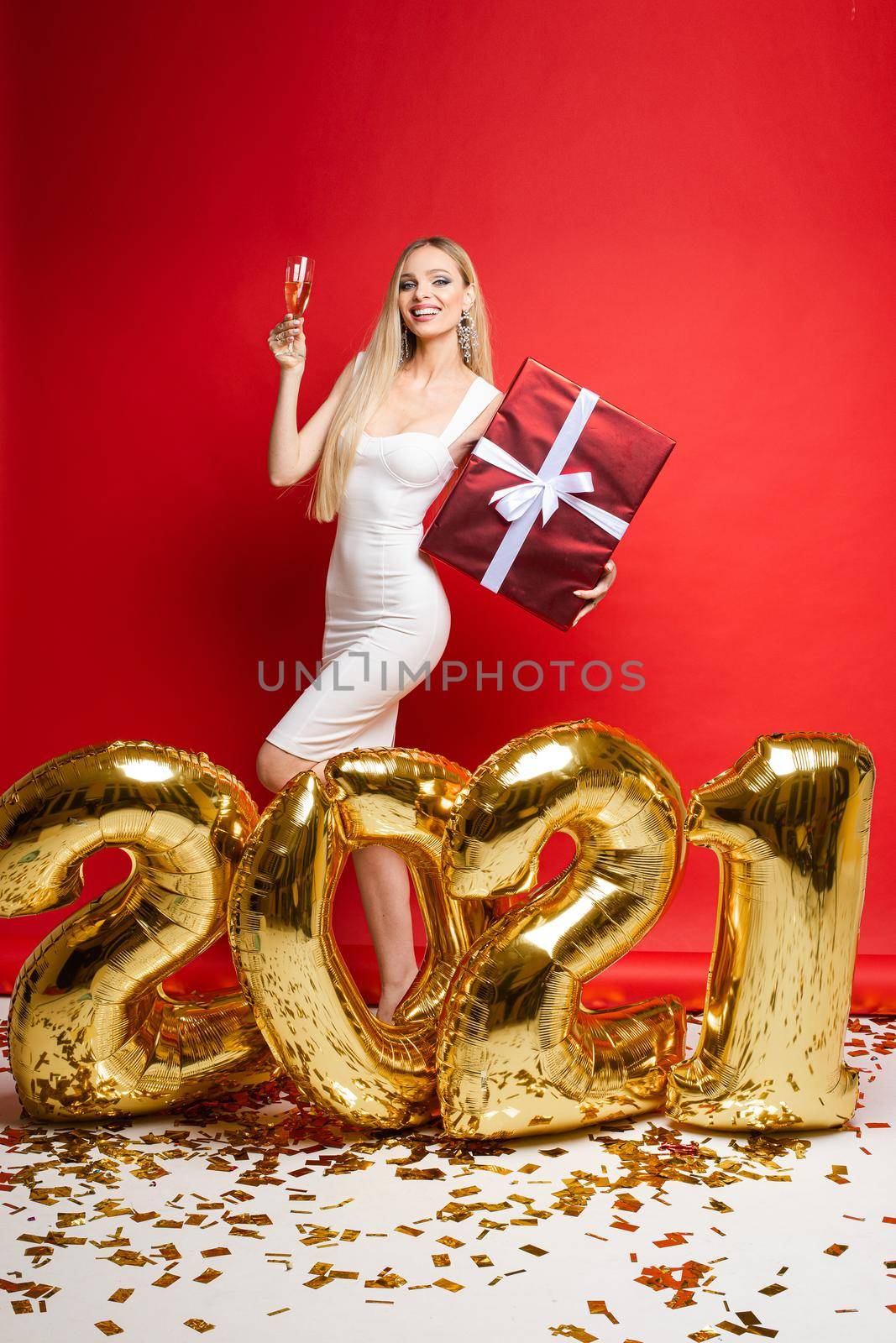 New year xmas celebrating, smiling girl with champagne, gift, golden balloons in 2021 shape, confetti on red background by StudioLucky