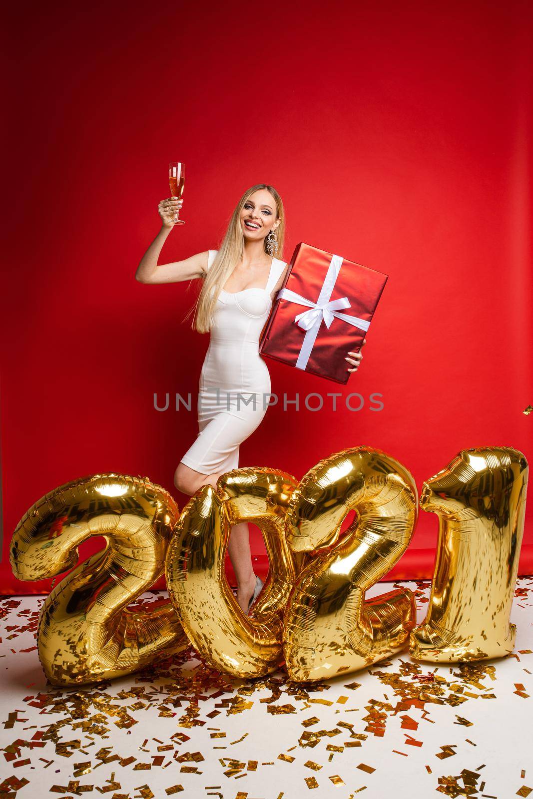 pretty woman in elegant white dress near big gold baloons with numbers 2021 and holds a big christmas gift and champagne by StudioLucky