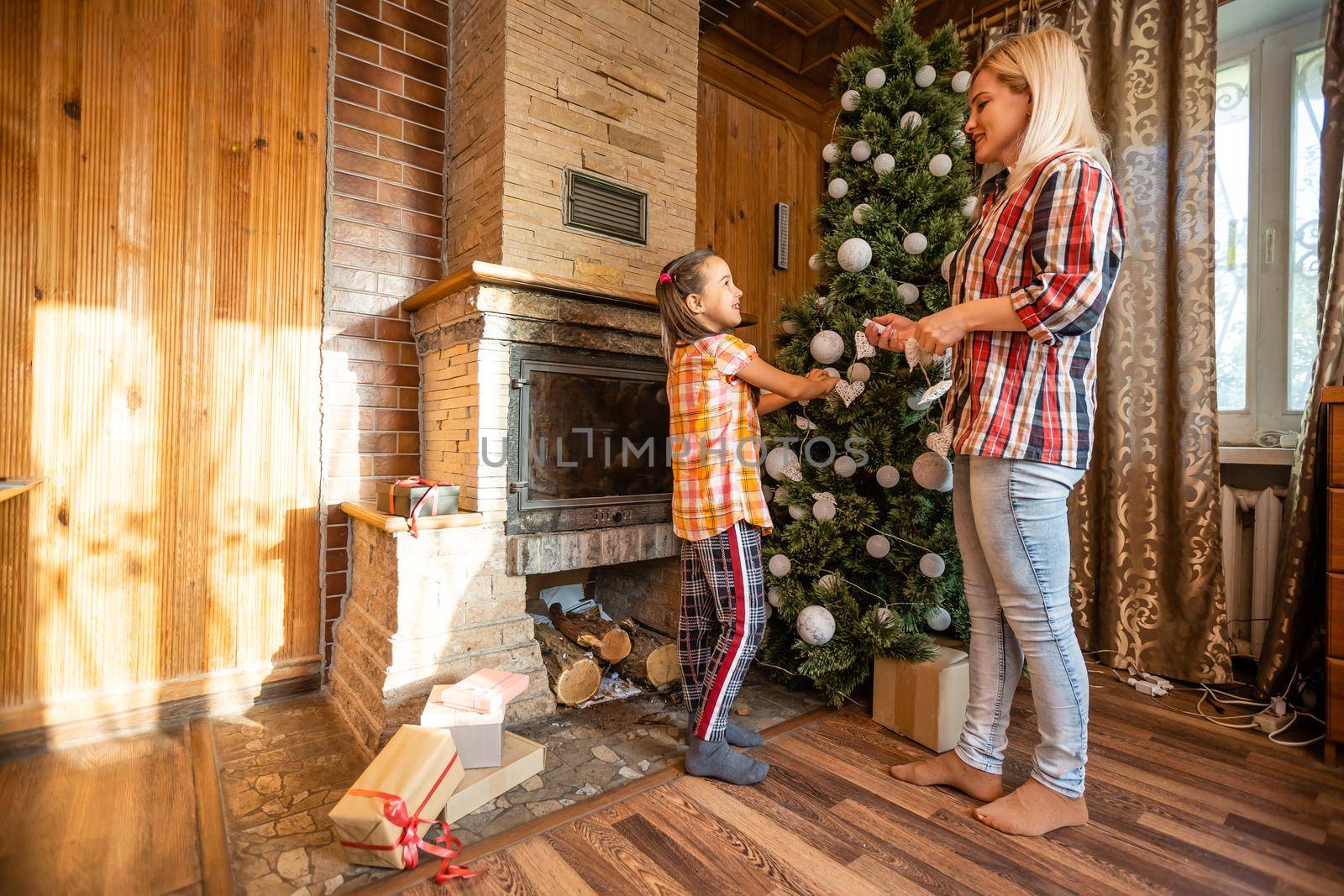 Mother holds a little daughter at the Christmas tree on a festive evening in a rustic wooden house, New Year, winter time