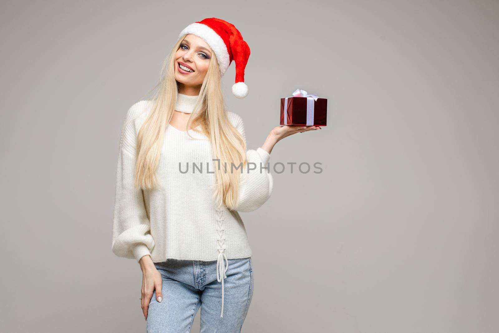 Stock photo of attractive blonde girl in red Santa hat and white sweater holding in hand a red Christmas present. Isolated on white.