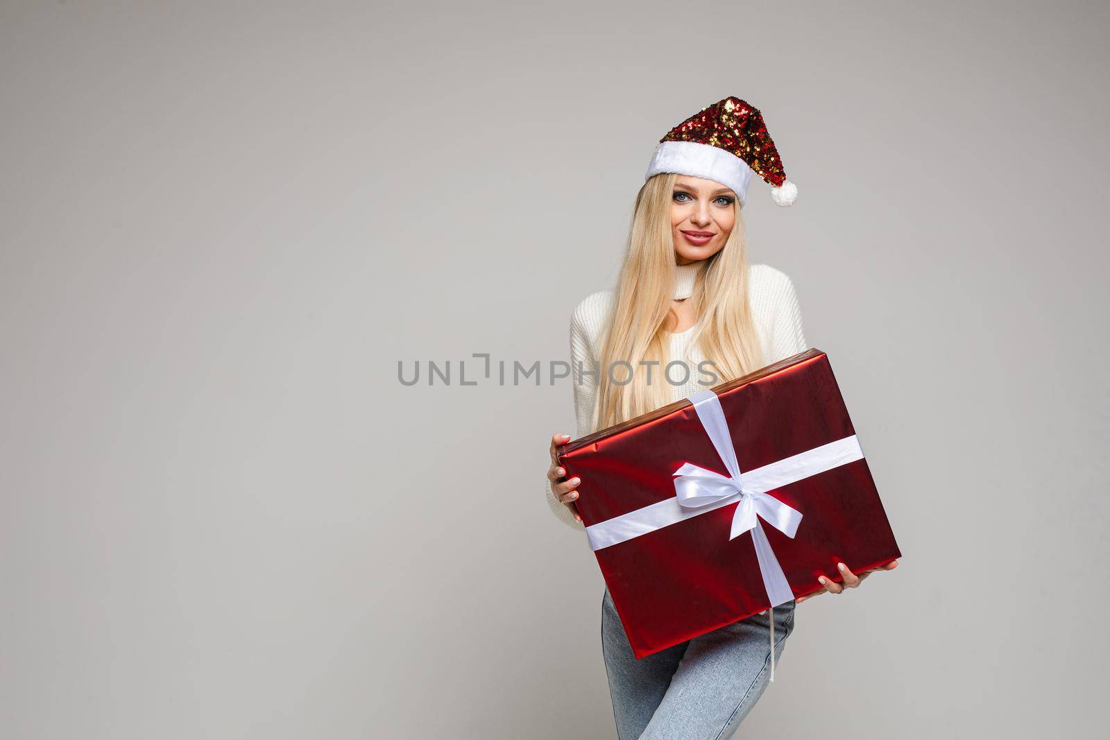 handsome caucasian woman with blonde hair and pretty face in warm sweater, jaens and christmas hat holds a big red box with a gift