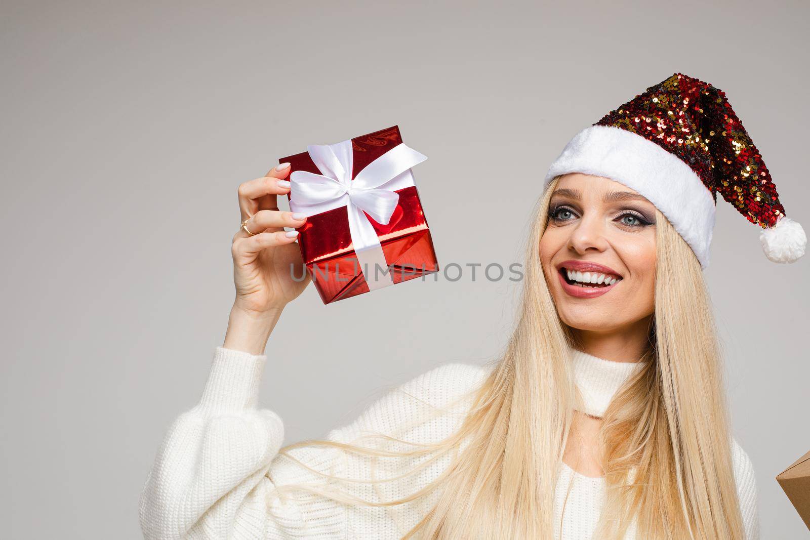 Portrait of pretty smiling woman holding gift box while looking at camera, isolated on grey background. New Year holiday concept