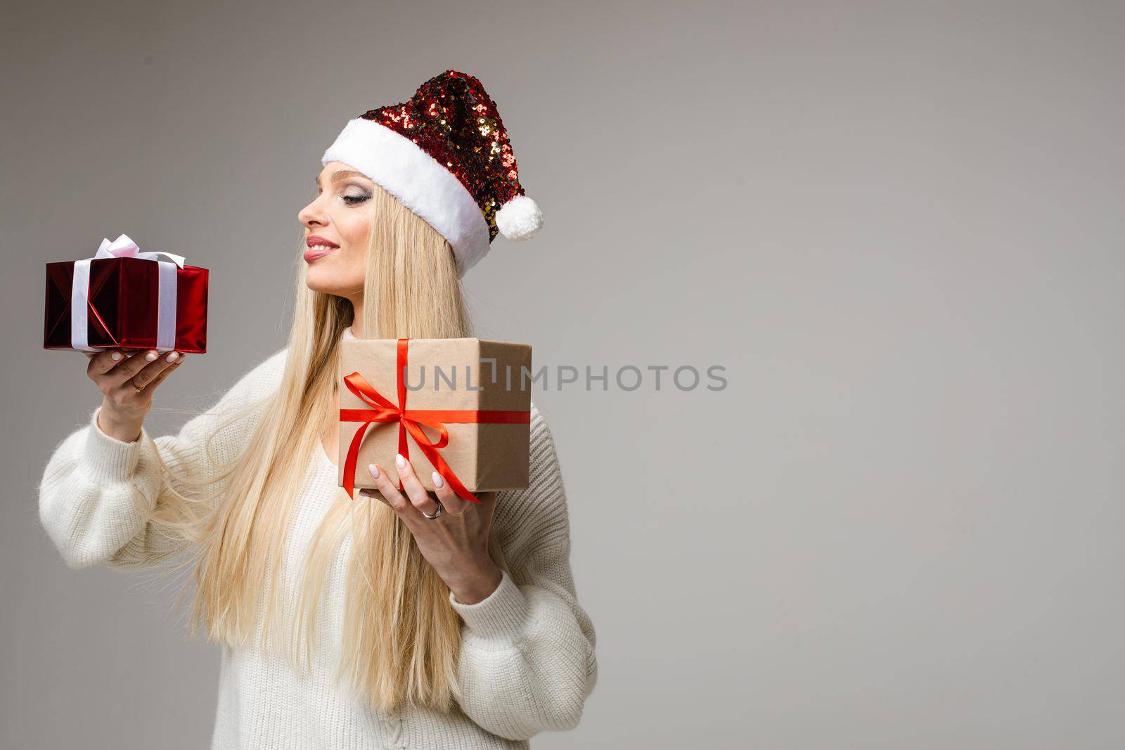 Stock photo of attractive blonde girl in red sparkling Santa hat and white sweater holding two Christmas presents and smiling over grey background. Copy space.