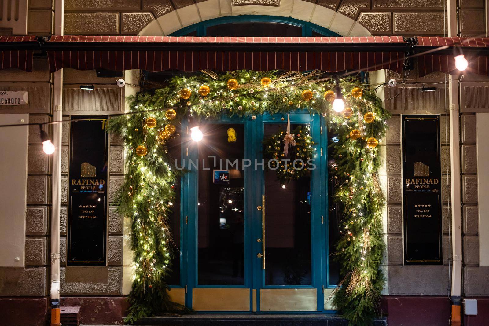 Stylish christmas tree with red berries, wreath on doors and wooden sleigh at front of store at holiday market in city street. Christmas street decor. Space for text. Rustic decoration.