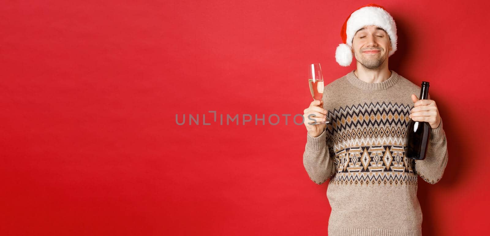 Concept of winter holidays, christmas and celebration. Image of pleased smiling man in santa hat and sweater, drinking on new year, holding bottle of champagne and filled glass, red background by Benzoix