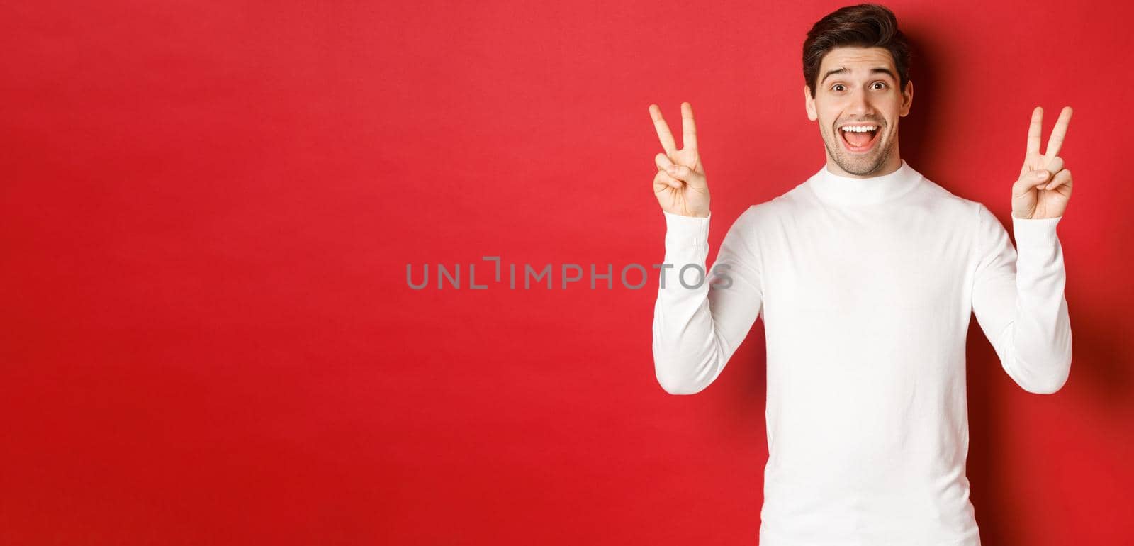 Concept of winter holidays, christmas and lifestyle. Handsome funny guy in white sweater, showing peace signs and smiling happy, standing against red background.