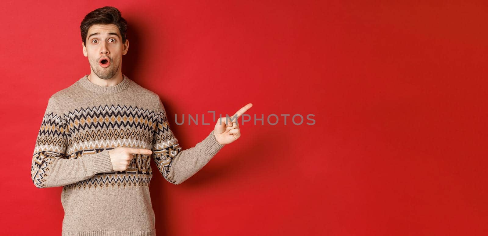 Concept of christmas celebration, winter holidays and lifestyle. Amazed handsome guy in xmas sweater pointing fingers right at logo, showing advertisement, standing over red background.