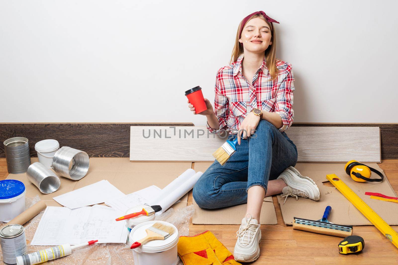 Dreamy girl painter relaxing on floor with cup of coffee. House remodeling and interior renovation concept with copy space. Young woman in red checkered shirt and jeans planning to redesign her home.