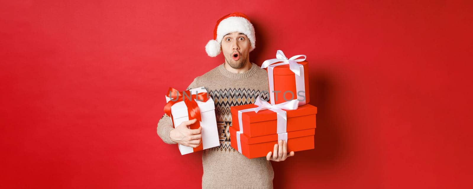 Concept of winter holidays, new year and celebration. Image of surprised handsome guy in sweater and santa hat, receiving gifts, holding boxes with presents and looking amazed by Benzoix